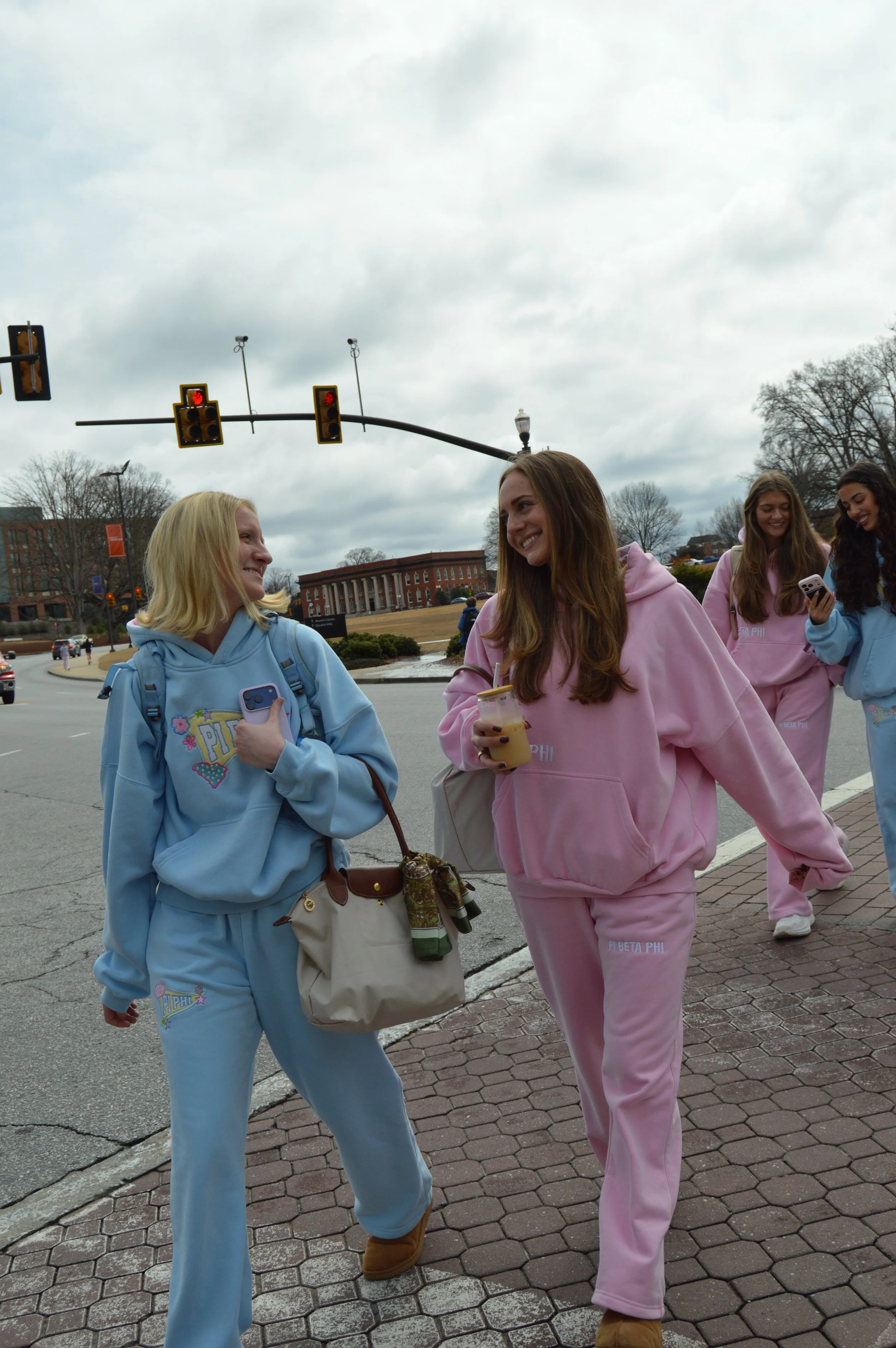 Group of young women walking on the sidewalk, wearing pastel-colored hoodies and sweatpants, smiling, with traffic lights overhead on a cloudy day.
