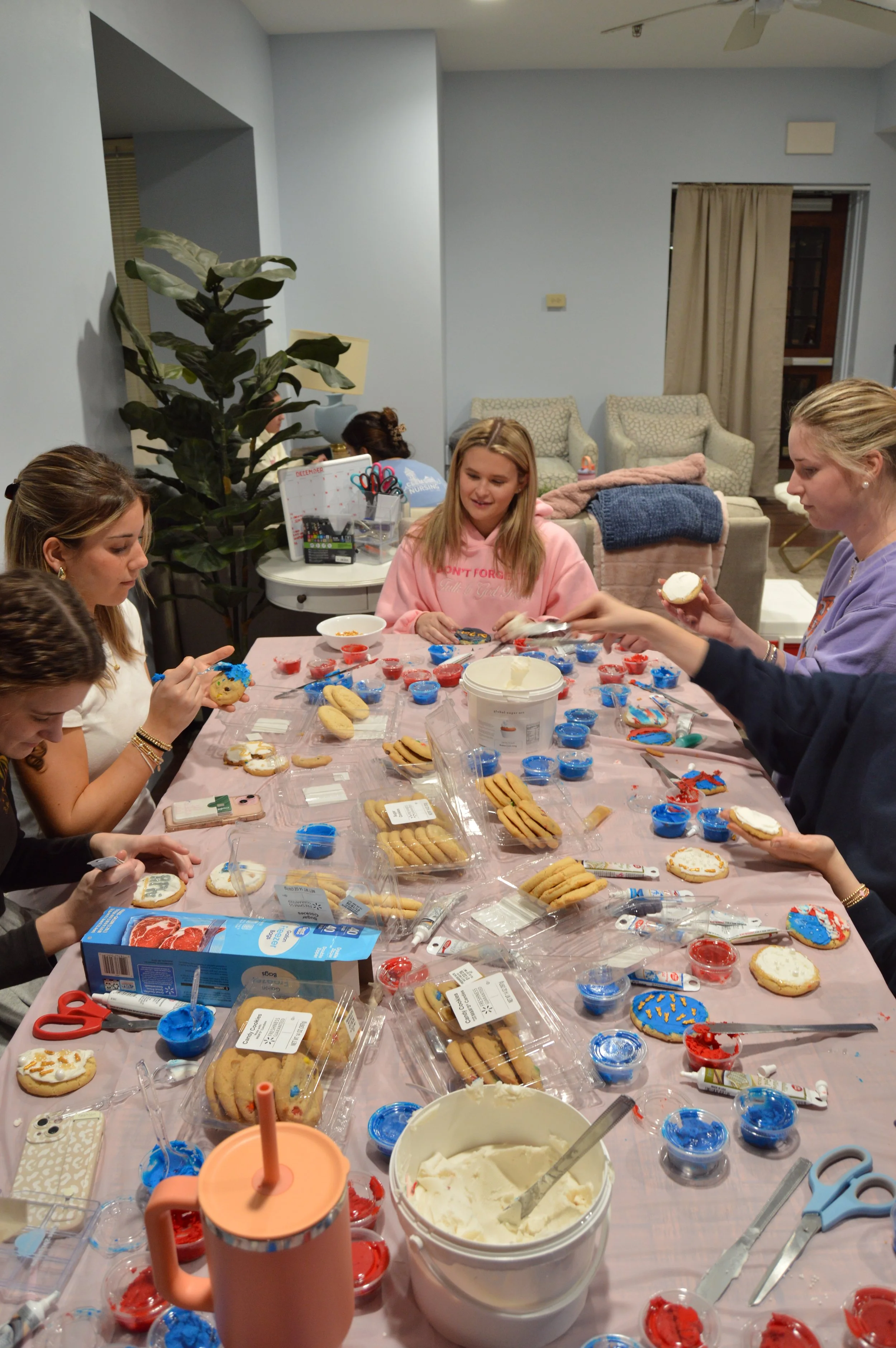 A group of six young women gathered around a long table decorated with various decorated cookies, frosting, and candy, decorating cookies together in a cozy indoor setting.