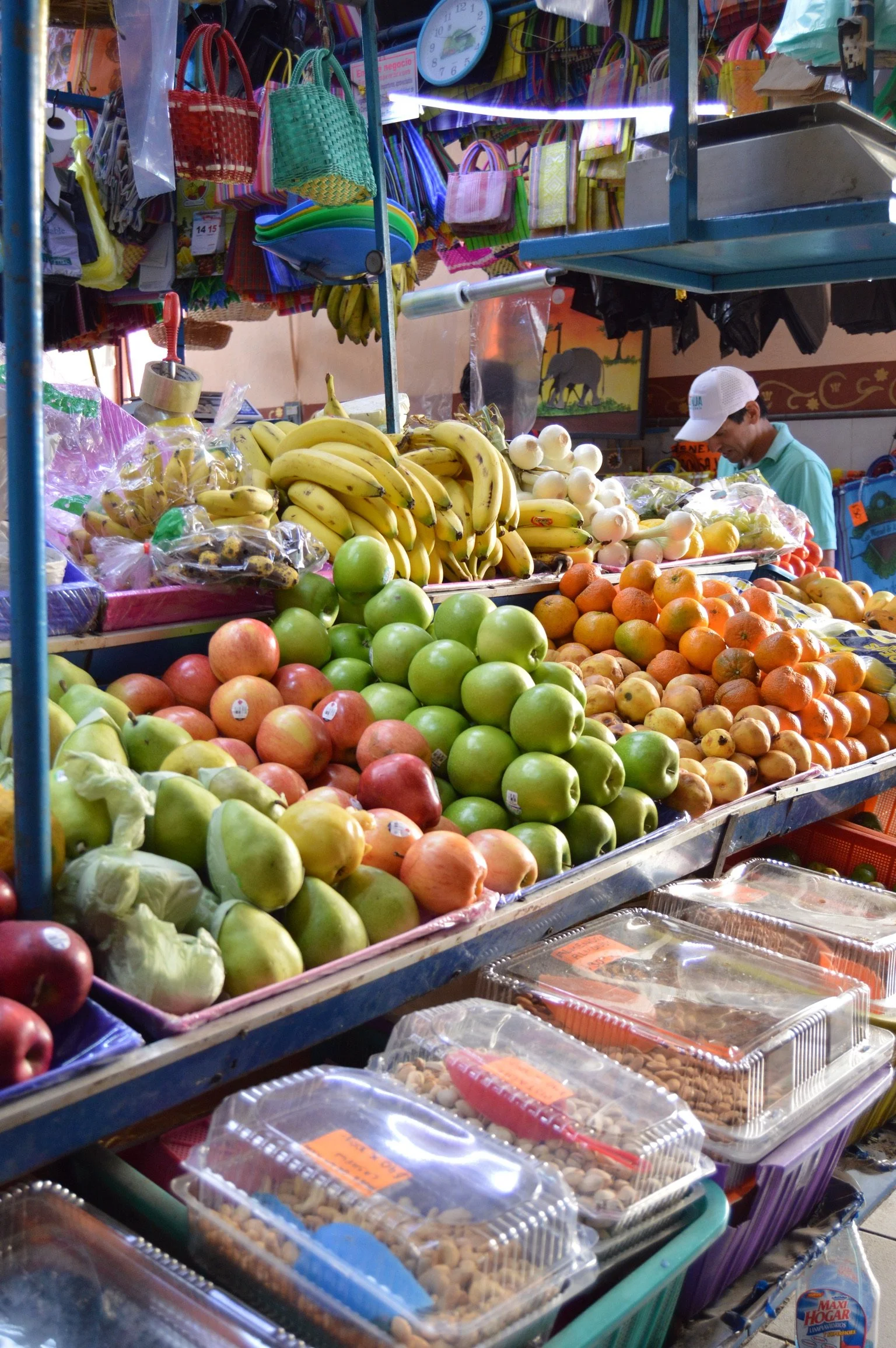 A colorful fruit stand with bananas, apples, oranges, and other fruits, along with packaged nuts, in an indoor market with hanging bags and a man shopping.
