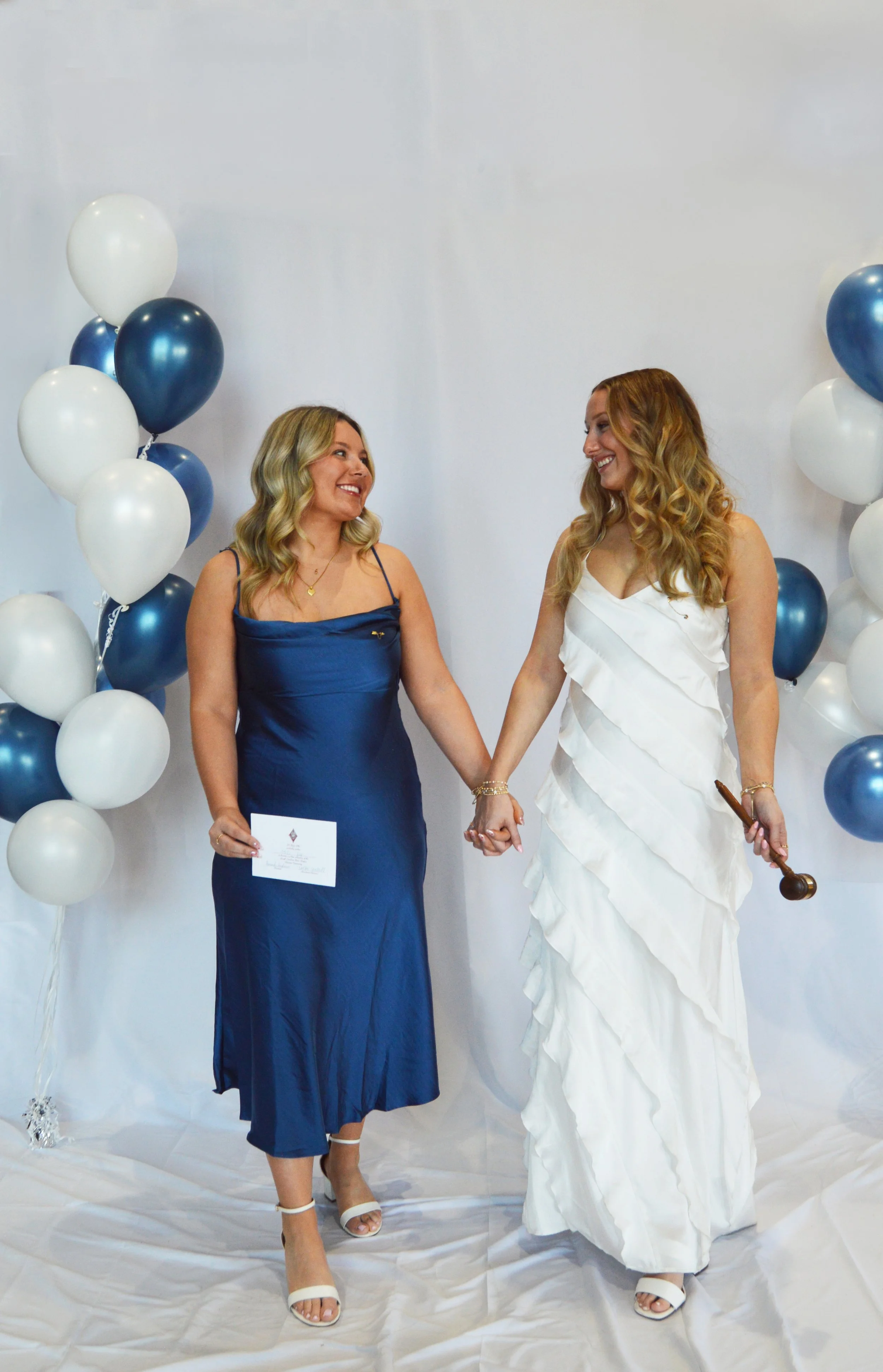 Two women, one in a blue dress and the other in a white dress, holding hands and smiling at each other in front of decorated balloons for a celebration or wedding.