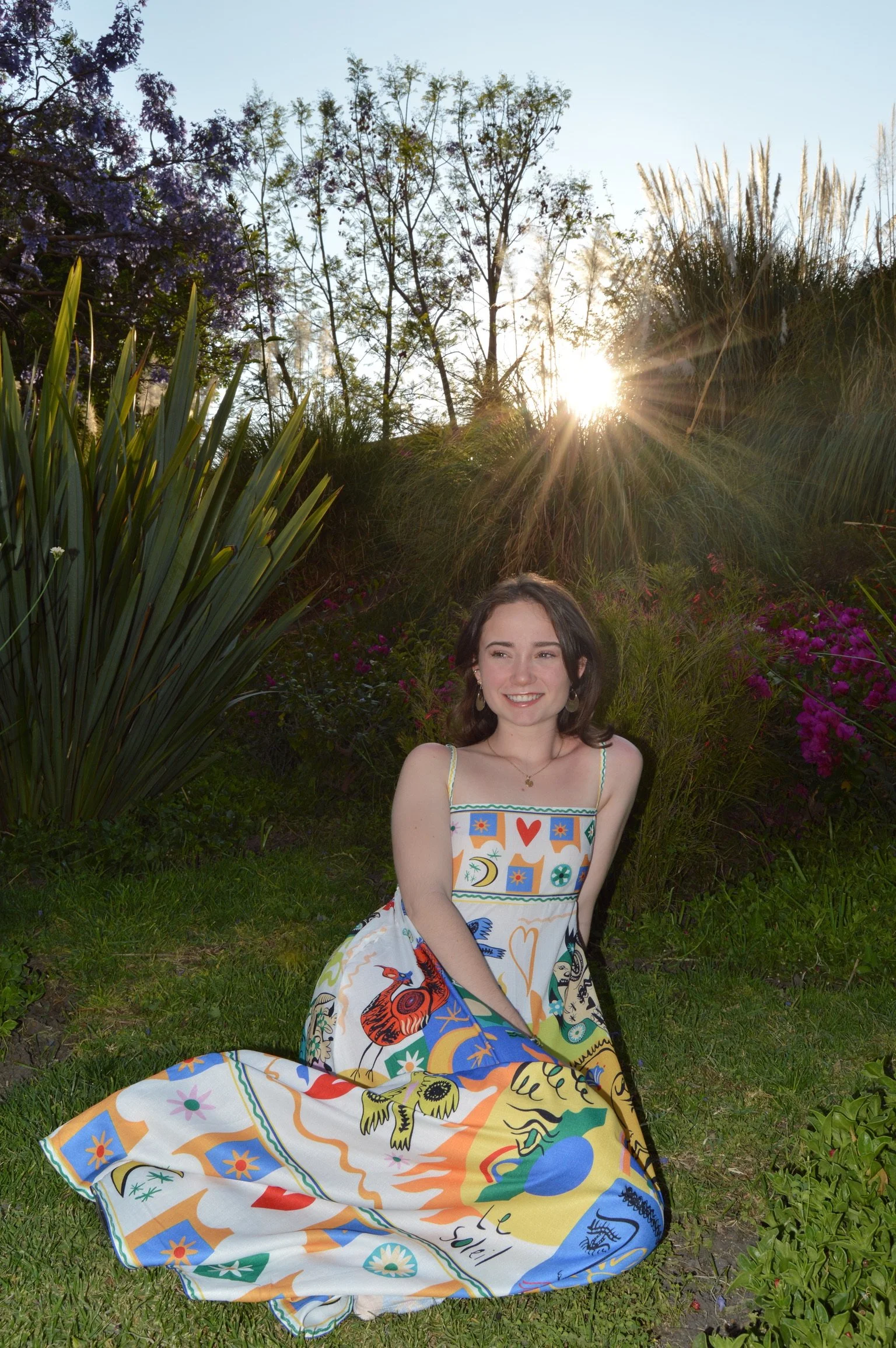 A young woman in a colorful, patterned dress sits on grass in a garden with purple flowers and tall greenery, with sunlight shining through trees in the background.