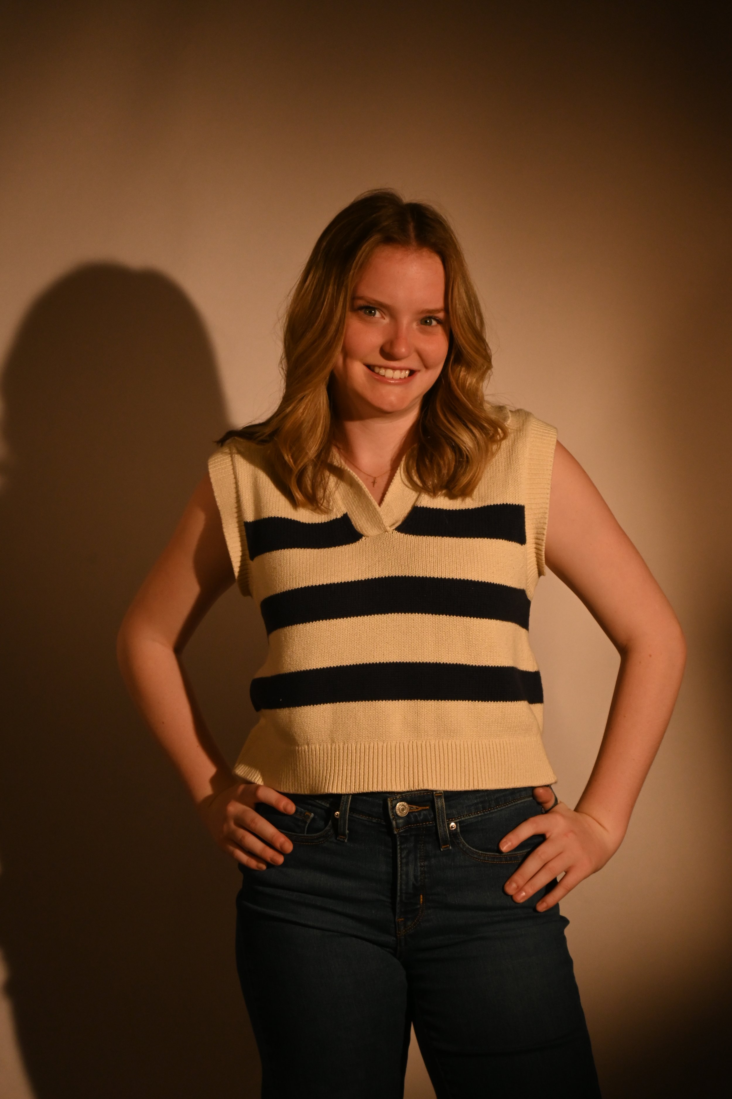 A young woman with shoulder-length blonde hair, smiling, wearing a sleeveless beige sweater with black horizontal stripes and dark jeans, standing against a plain background.