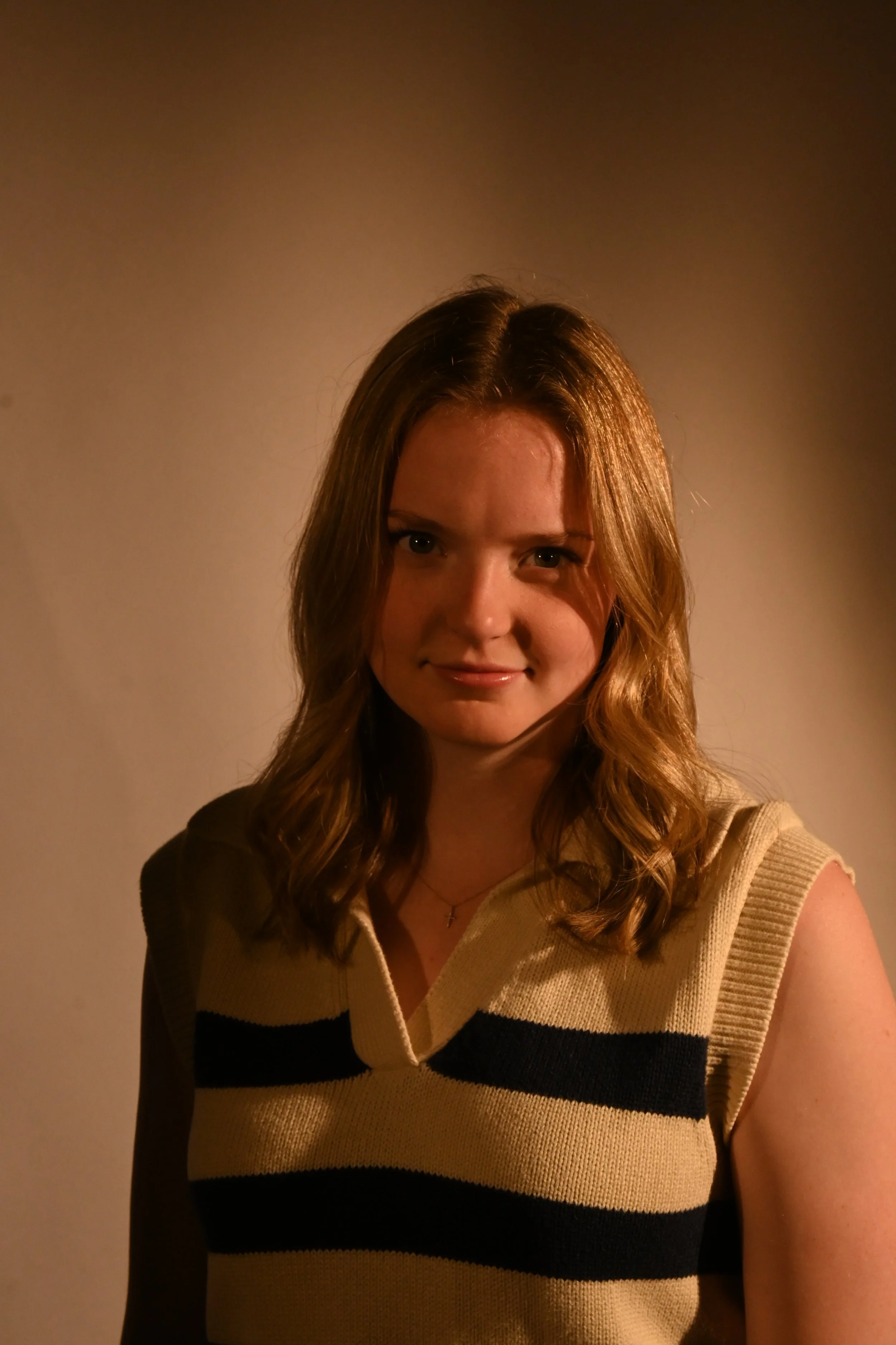 A young woman with shoulder-length brown hair, wearing a sleeveless beige sweater with black horizontal stripes, standing against a plain wall with warm lighting.