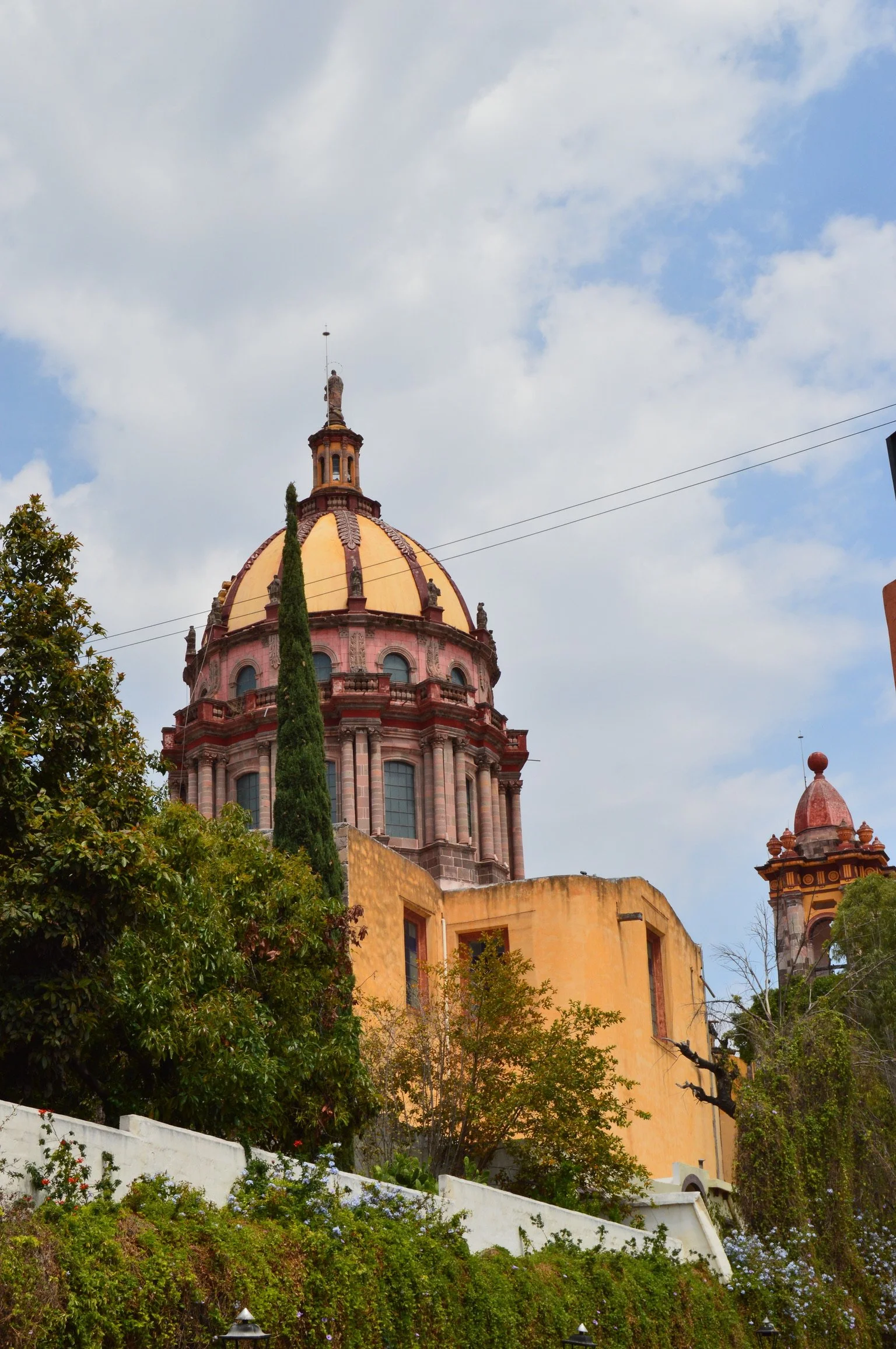 A historic building with a large domed roof, part of a church or cathedral, nestled among trees and greenery, with a partly cloudy sky in the background.