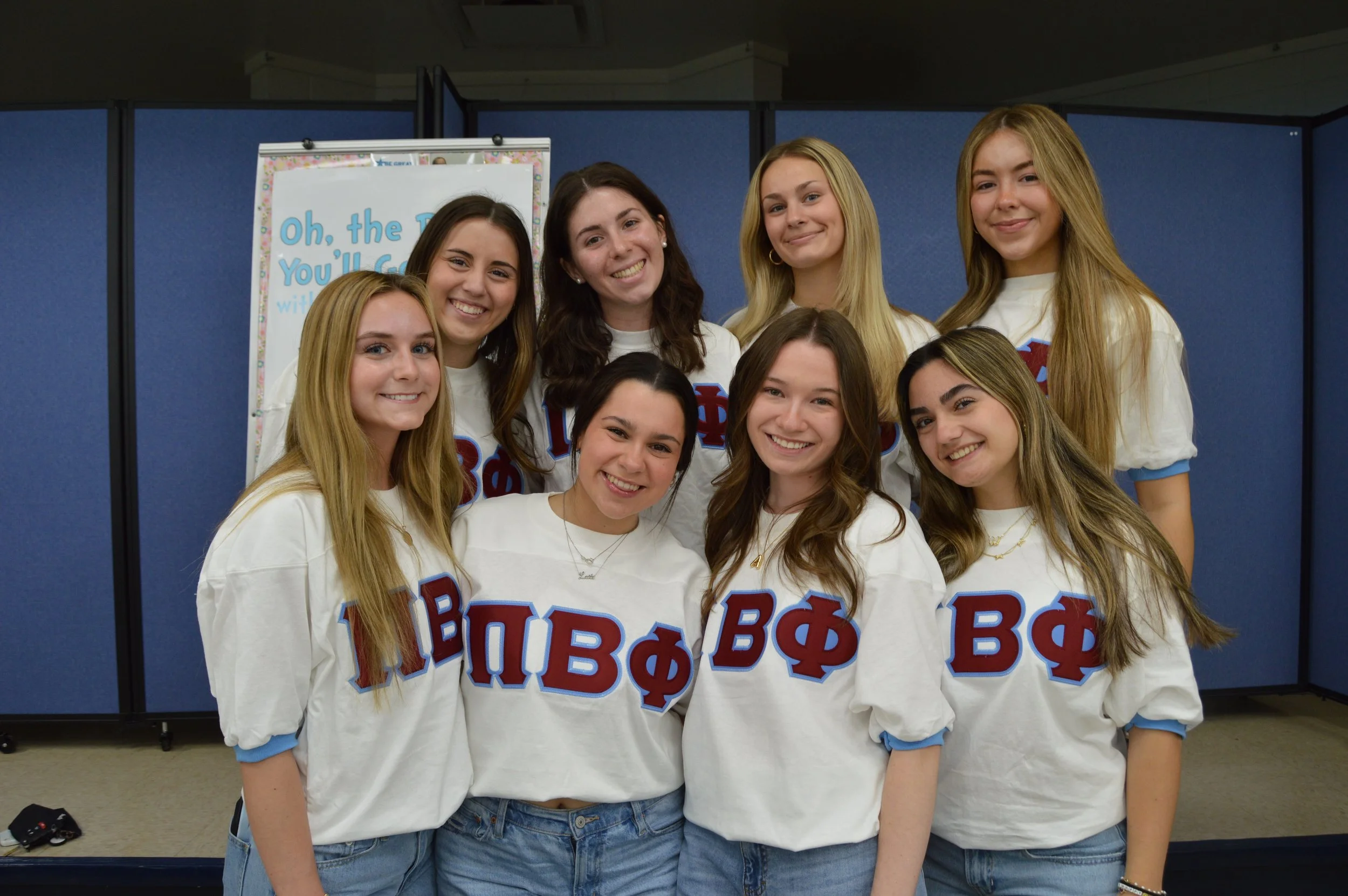 A group of nine young women smiling, standing together, wearing white sweatshirts with Greek letters, in front of a blue folding wall.