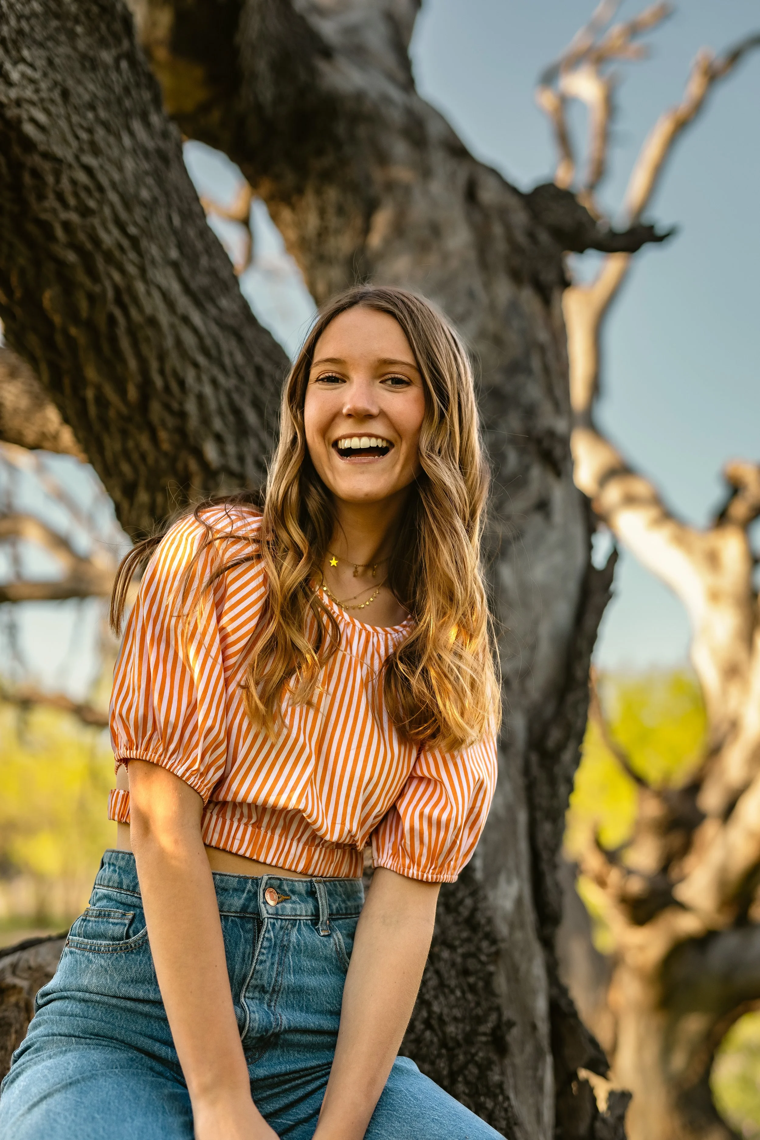 A young woman with long wavy hair, smiling and looking at the camera, standing outdoors next to a large, leafless tree with a blue sky in the background.