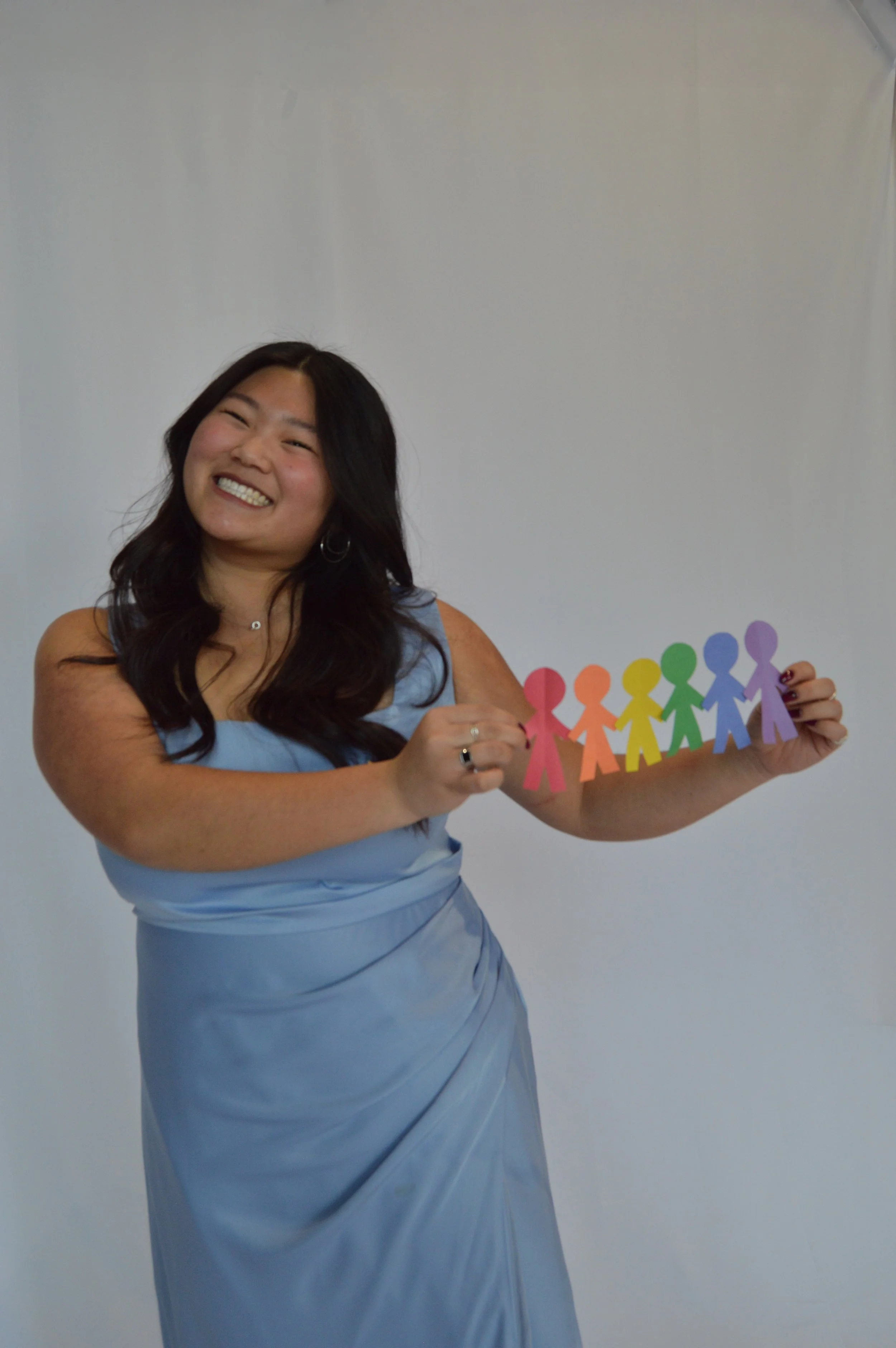 A woman in a light blue dress smiling and holding a colorful paper chain of paper figures.