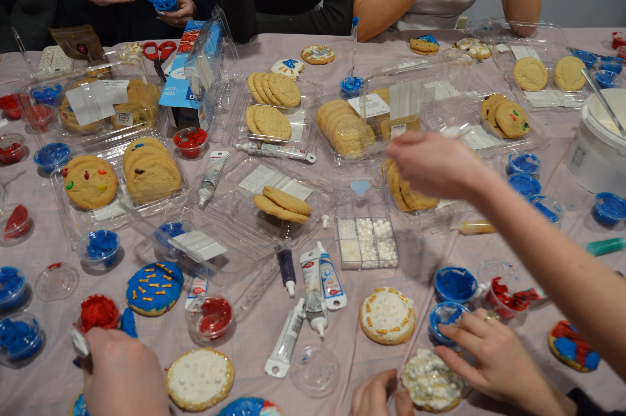 People decorating decorated sugar cookies with icing and sprinkles on a table with containers of colorful icing and cookie ingredients.