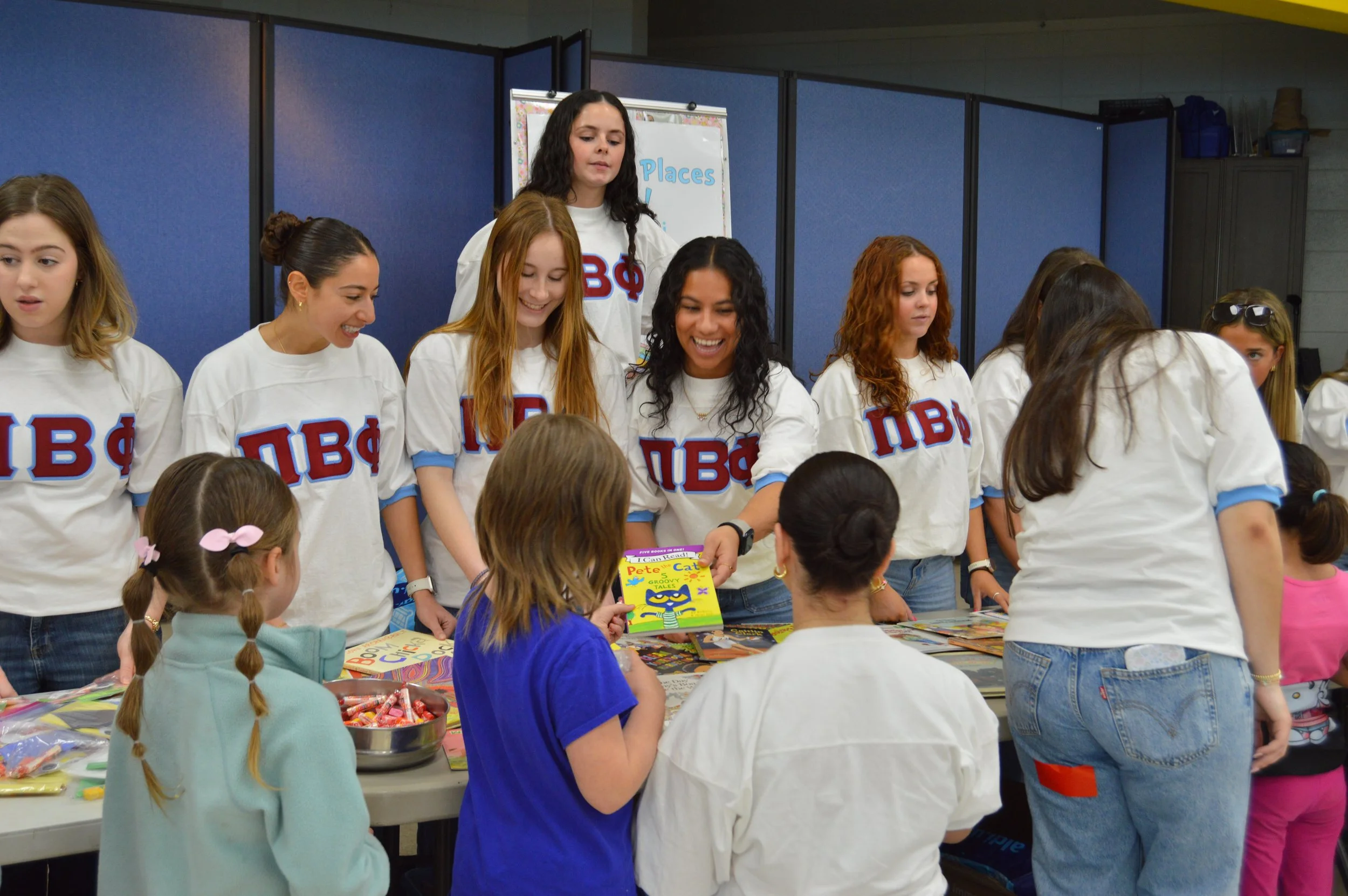 Group of young women in white BPhi shirts helping children at a table filled with books and toys.