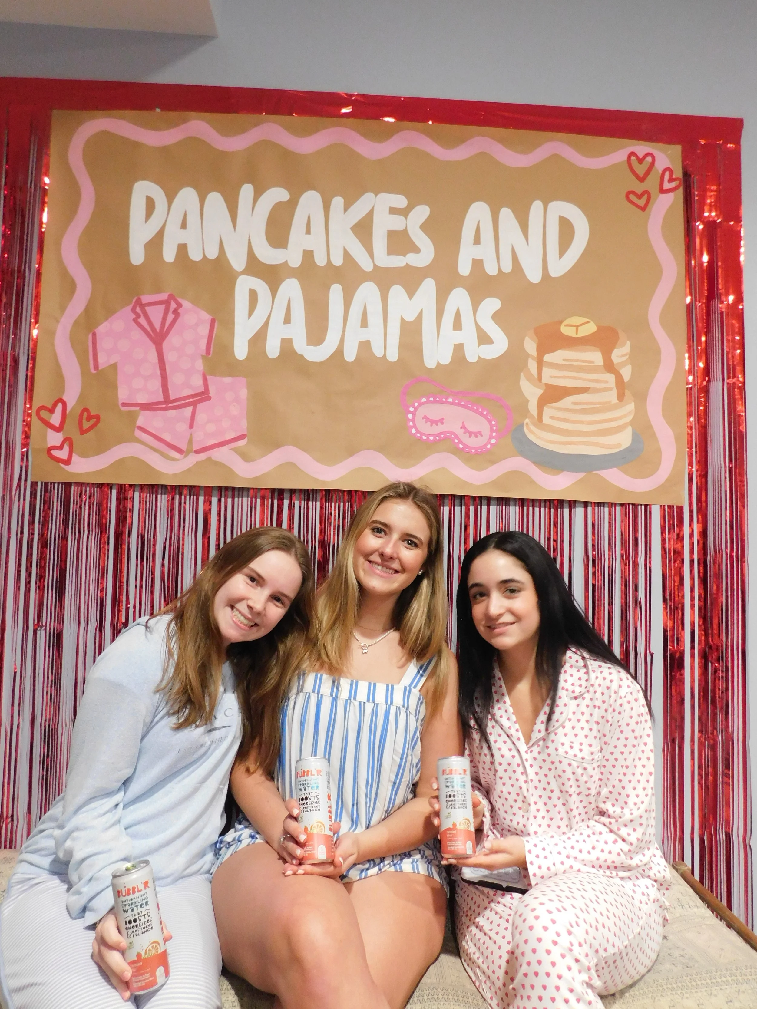 Three women sitting on the floor in pajamas, smiling, holding cans of sparkling water in front of a birthday-themed backdrop with a sign reading "Pancakes and Pajamas" that includes drawings of a pajama set, stacked pancakes, and a sleep mask.