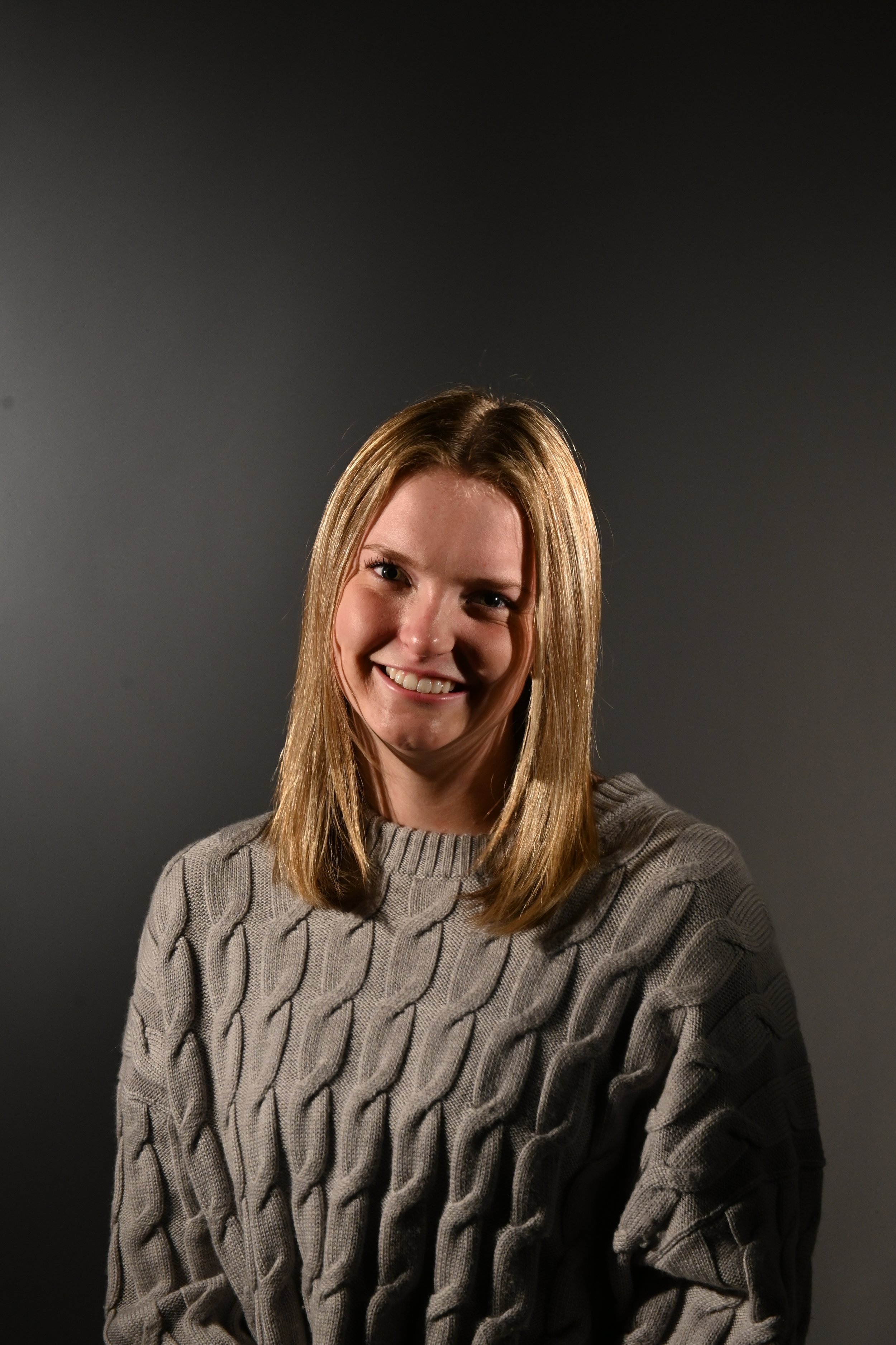 A young woman with shoulder-length blond hair smiling, wearing a beige knit sweater with a cable pattern, against a dark gray background.