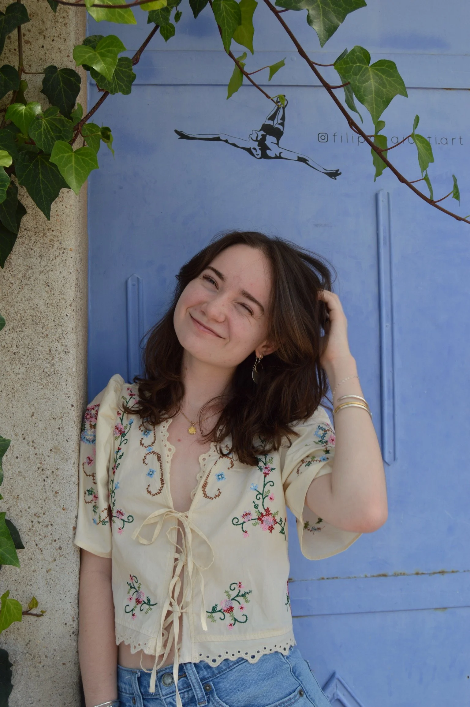 A young woman with shoulder-length brown hair, smiling and tilting her head slightly, wearing a cream-colored embroidered blouse and jeans, standing next to a blue door with a silhouette of an aerial performer above.