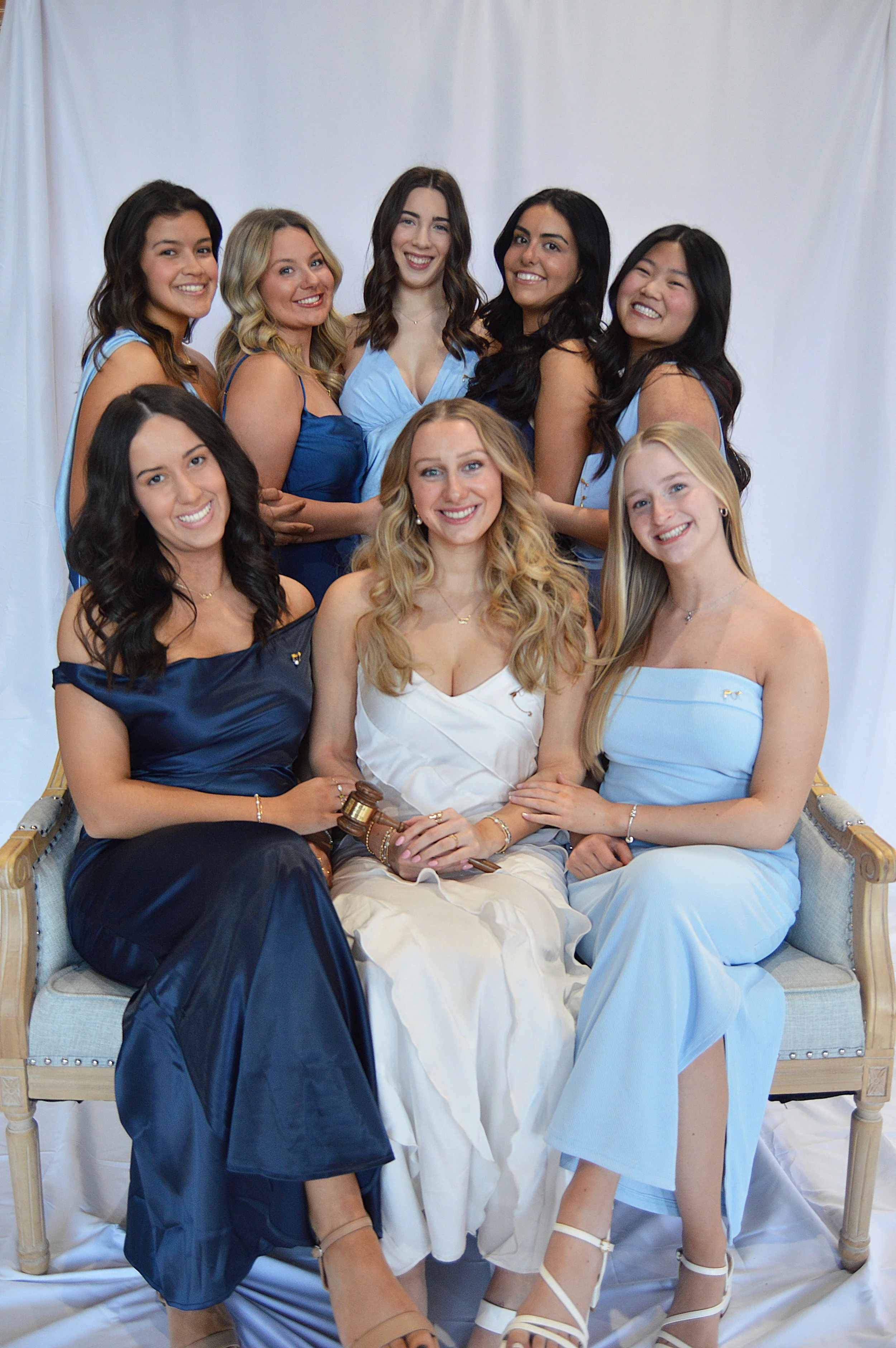 Group of nine young women in formal dresses, posing for a photo against a white backdrop. Some are seated, others are standing behind, all smiling.