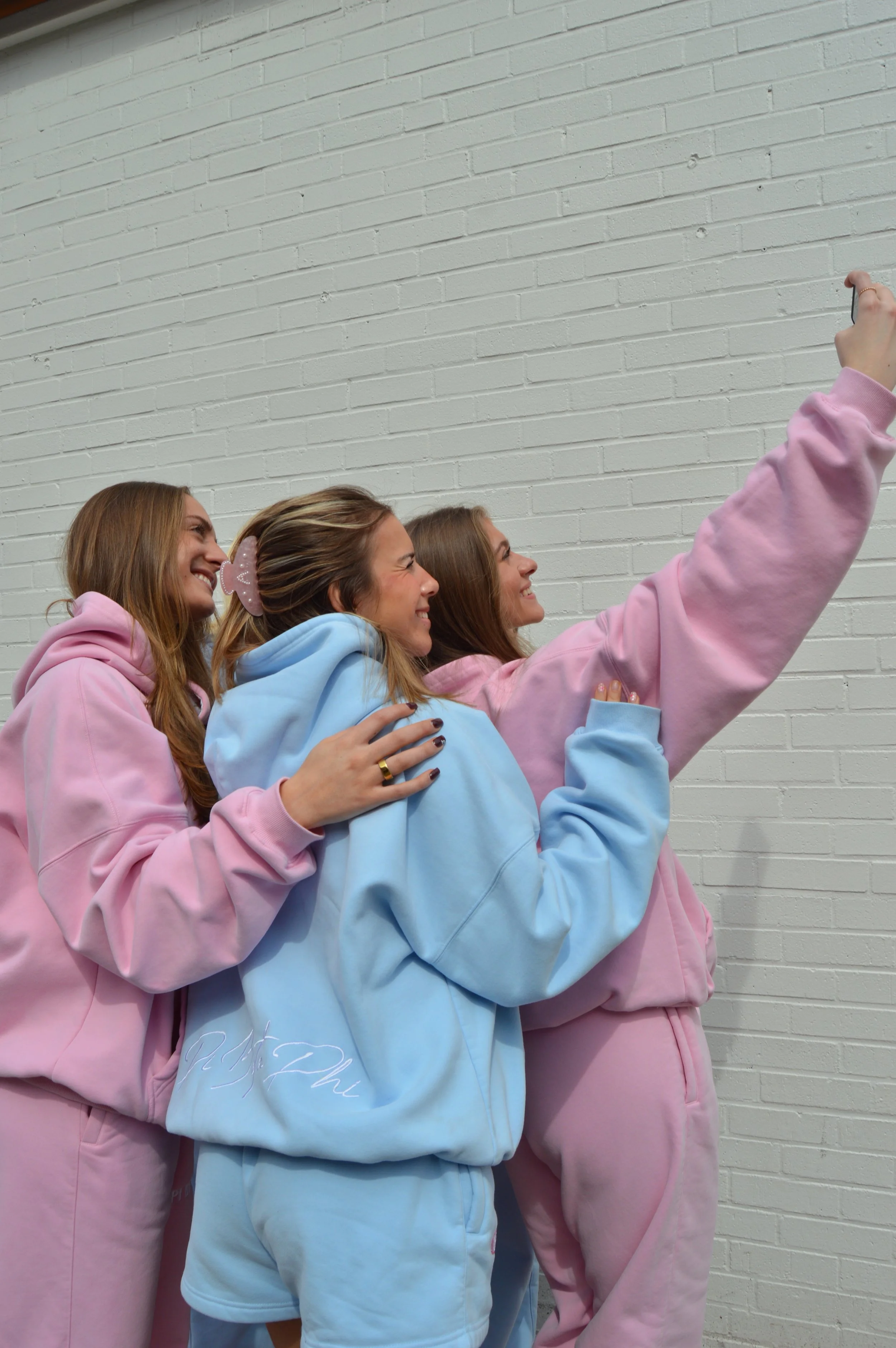 Four young women taking a selfie together against a white brick wall. They are dressed in pastel-colored hoodies and sweatshirts, smiling and posing for the photo.