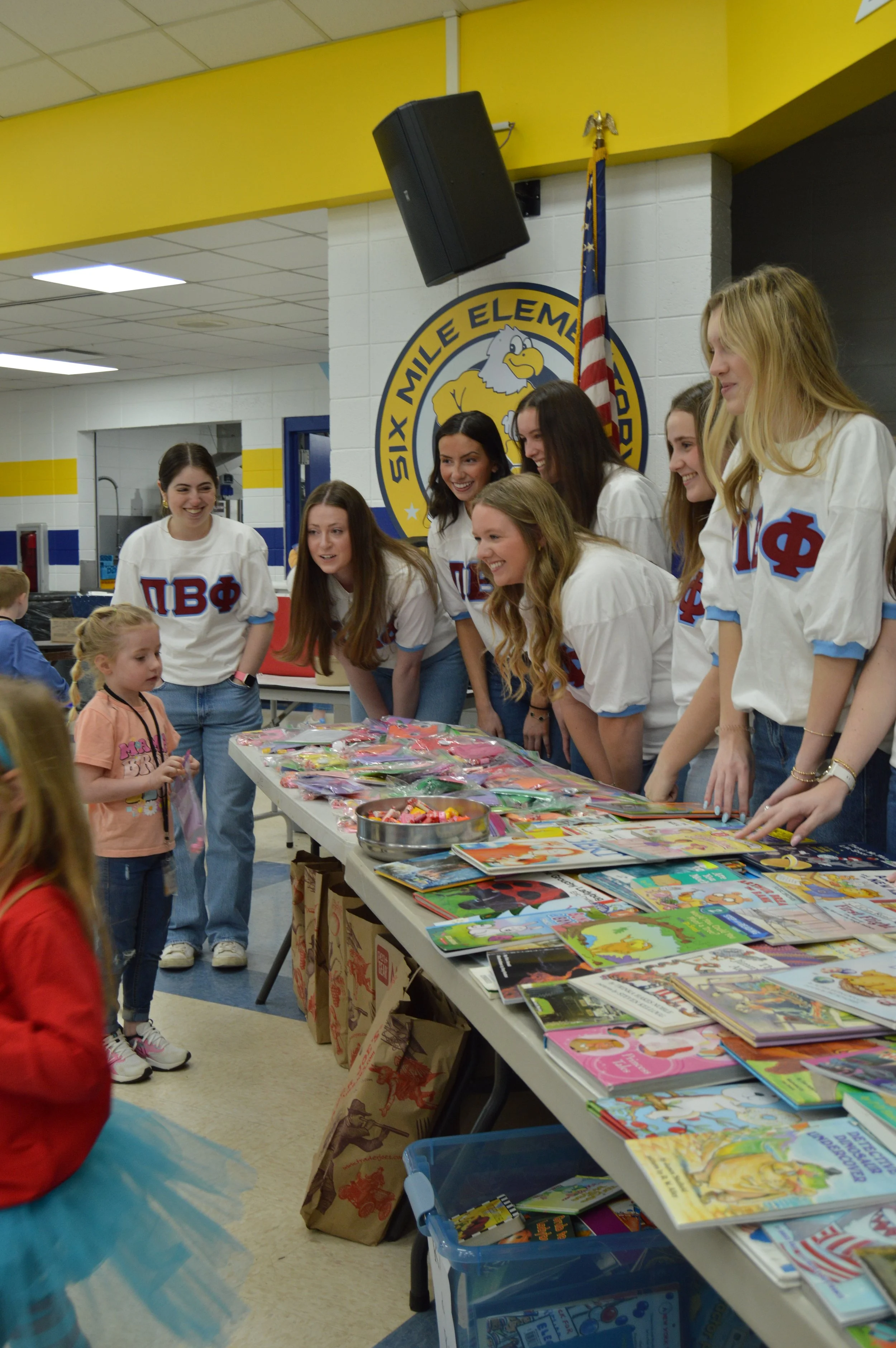 Students and children at a book fair or event in a school cafeteria, with a table full of books and candy, and a formation of smiling girls in sorority shirts. The school logo and American flag are visible in the background.