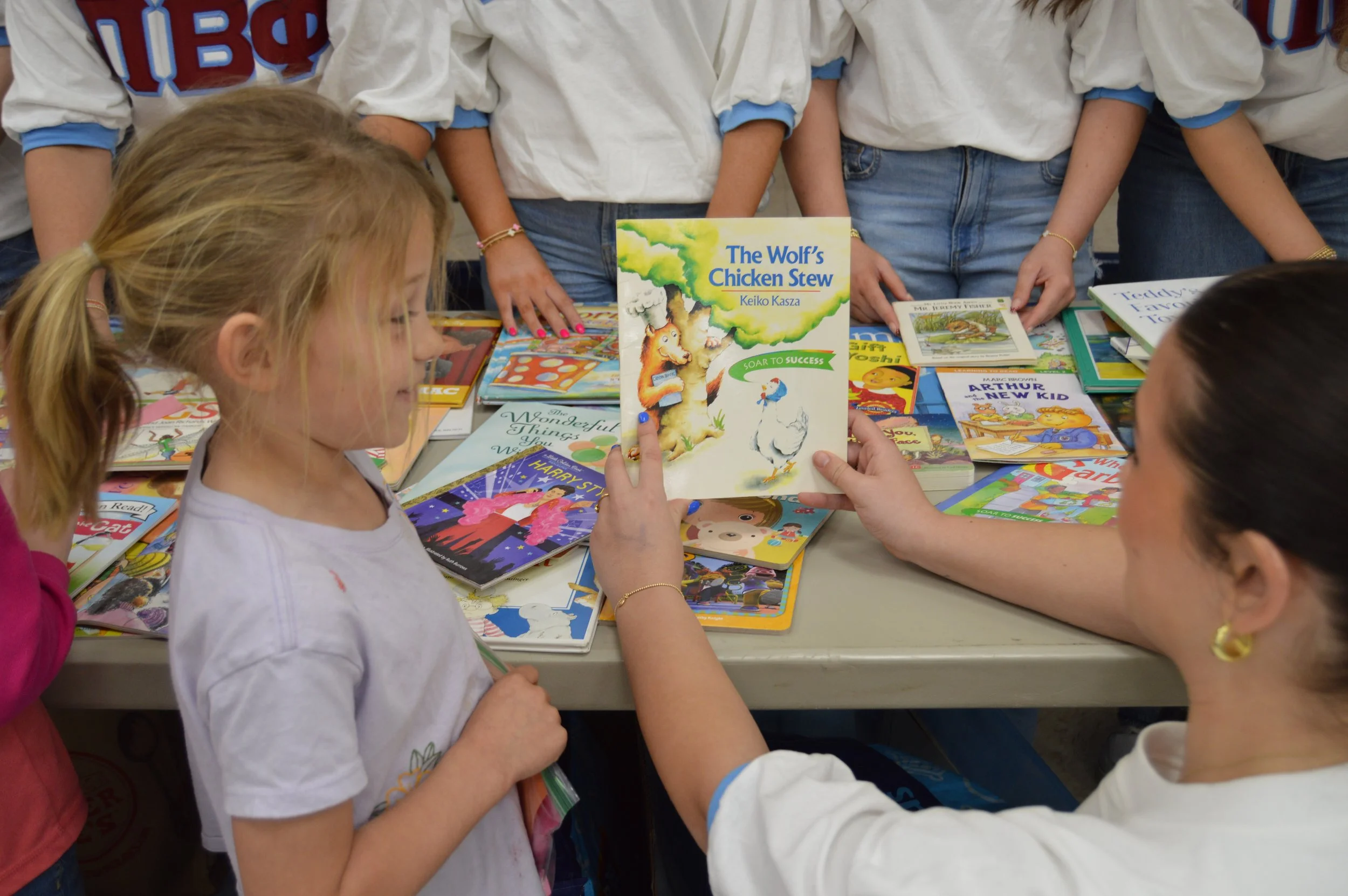 A woman holding a children's book titled 'The Wolf's Chicken Stew' shows it to a girl with blonde hair at a table covered with various colorful children's books, surrounded by other children in white shirts with blue accents.