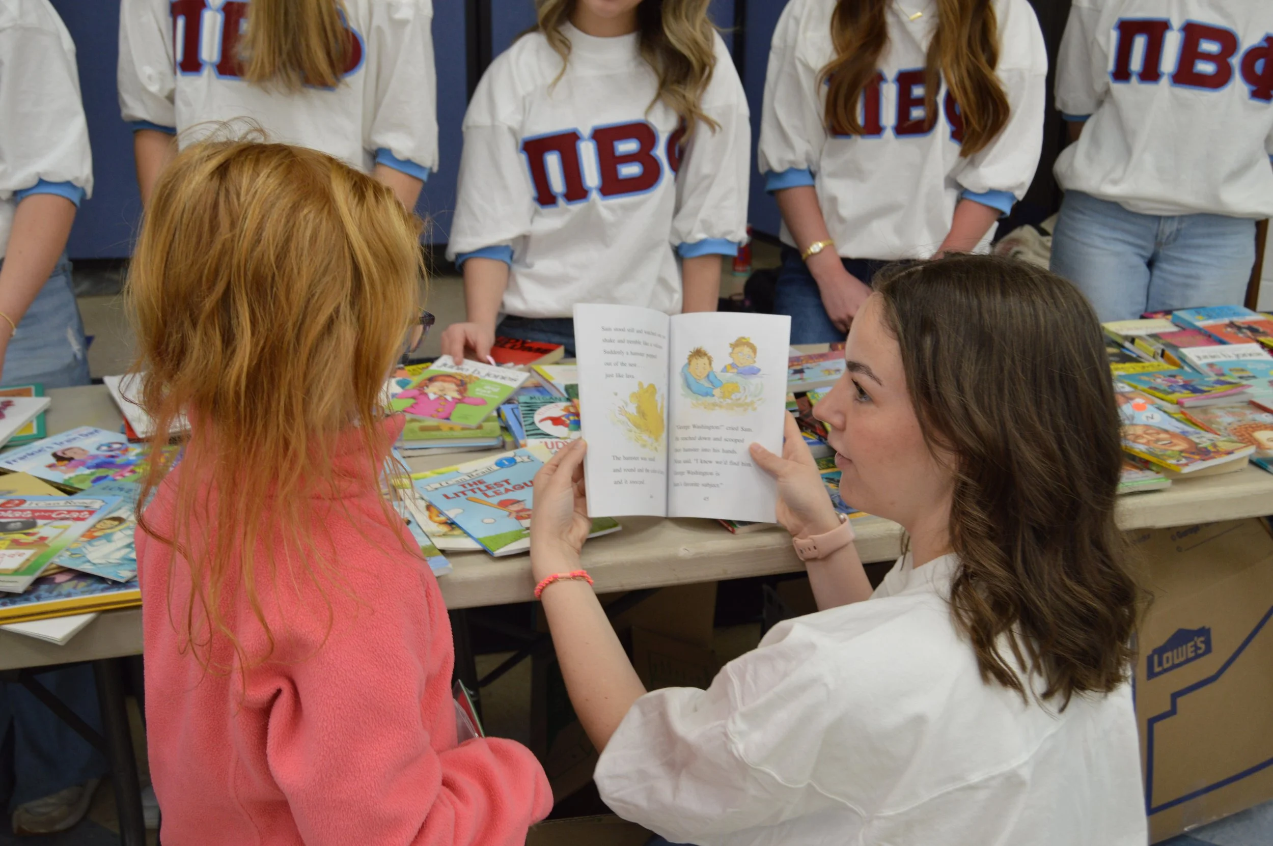 A woman showing a children's book to a young girl at a book sale event, with other books spread out on the table and several people standing in the background wearing matching shirts.