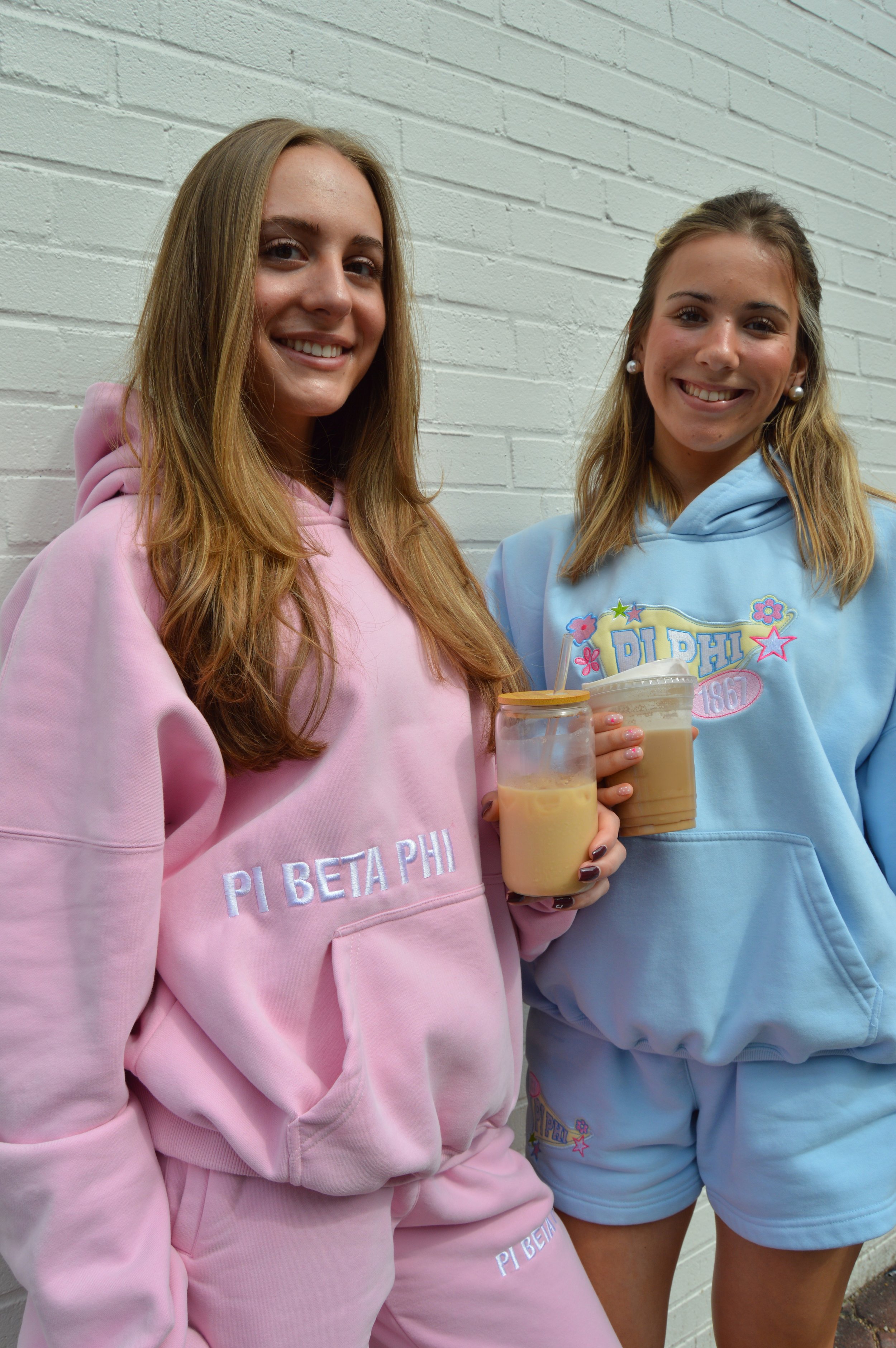Two young women smiling and holding bubble tea drinks, standing against a white brick wall. One is wearing a pink hoodie with 'PI BETA PHI' on it, and the other is wearing a light blue hoodie with a similar design.