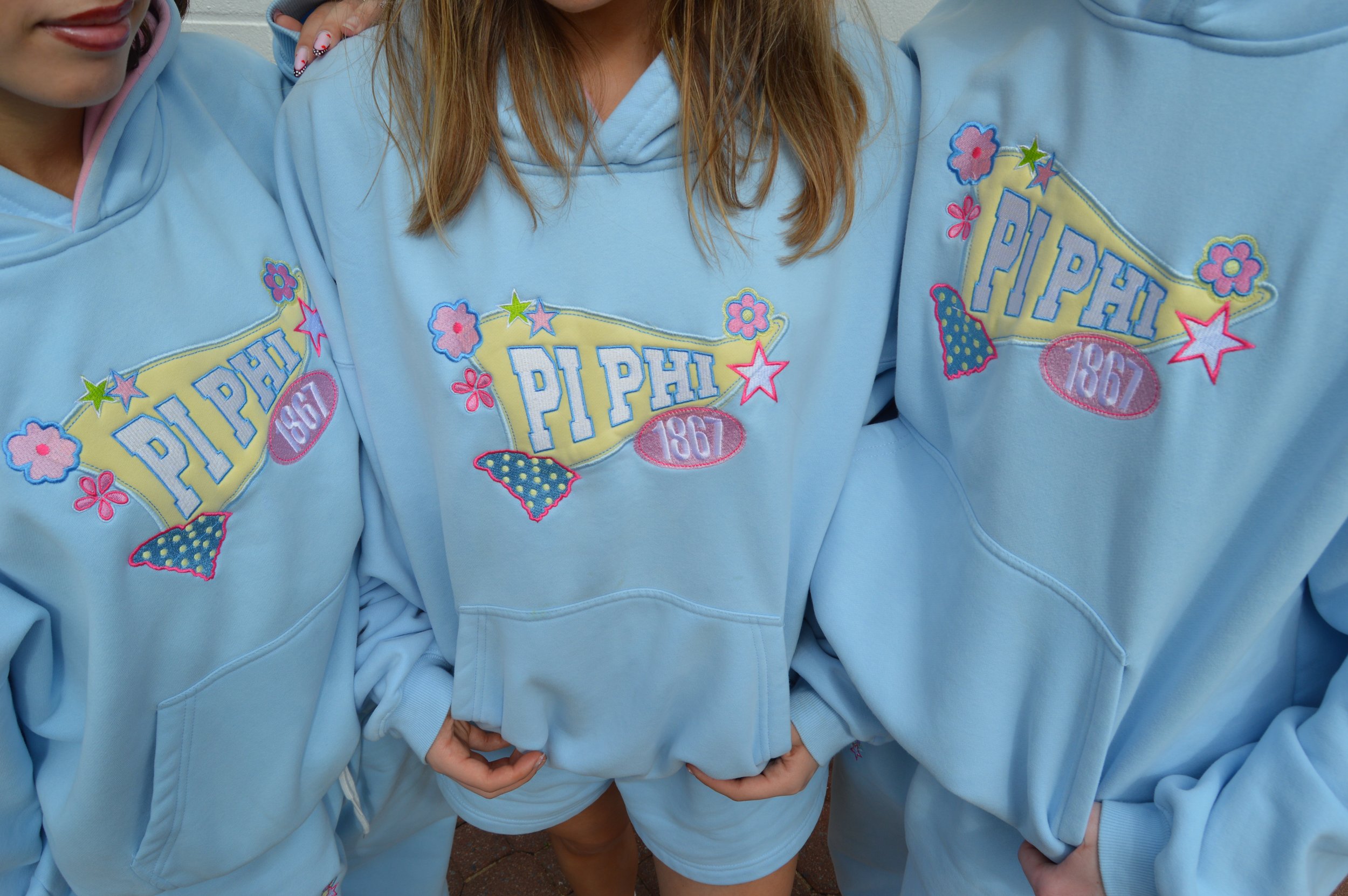 Close-up of three people wearing matching light blue sweatshirts with colorful embroidered patches that say "Piphi 1867" and decorated with flowers, stars, and a strawberry design.