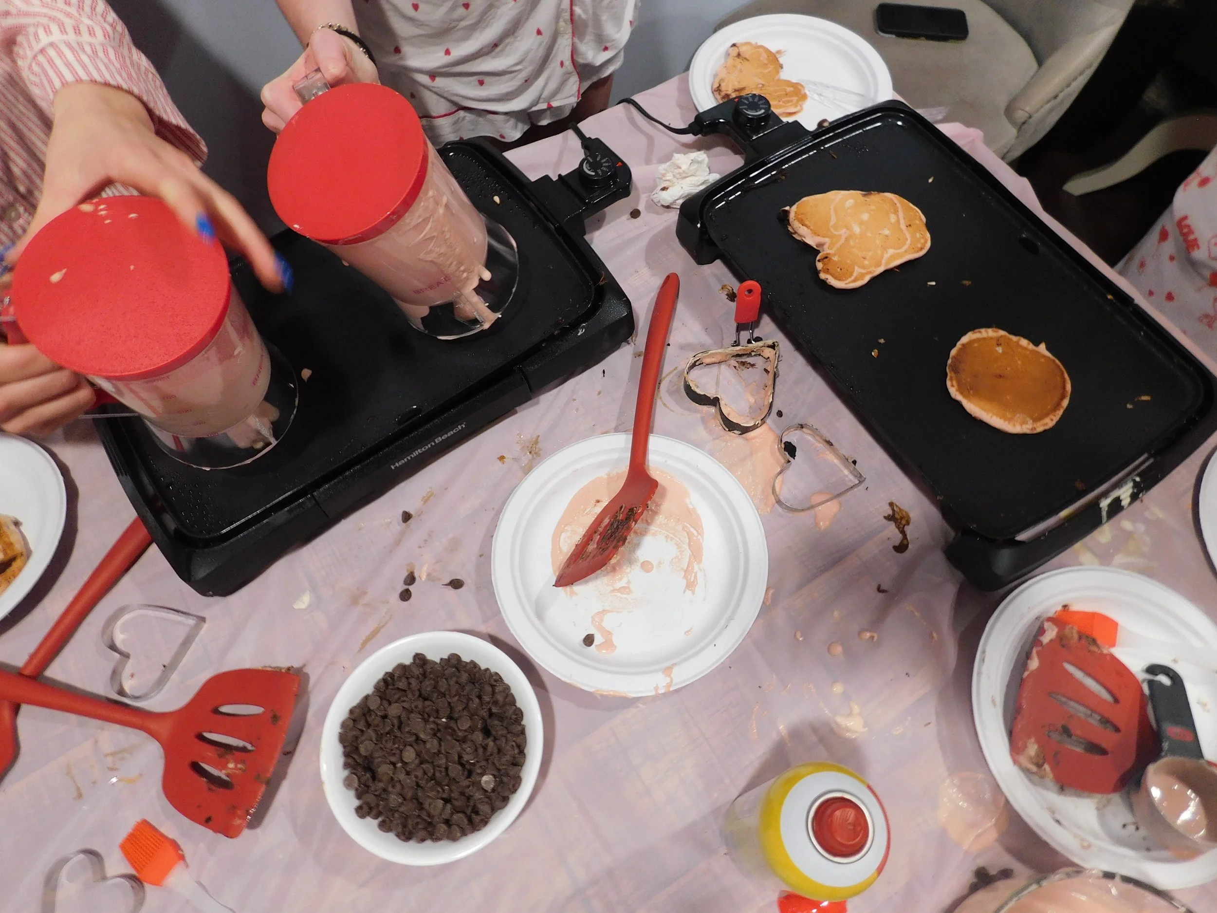 A messy table with pancake-making supplies, batter, chocolate chips, waffle maker, spatulas, and partially cooked pancakes. Hands are holding syrup bottles.