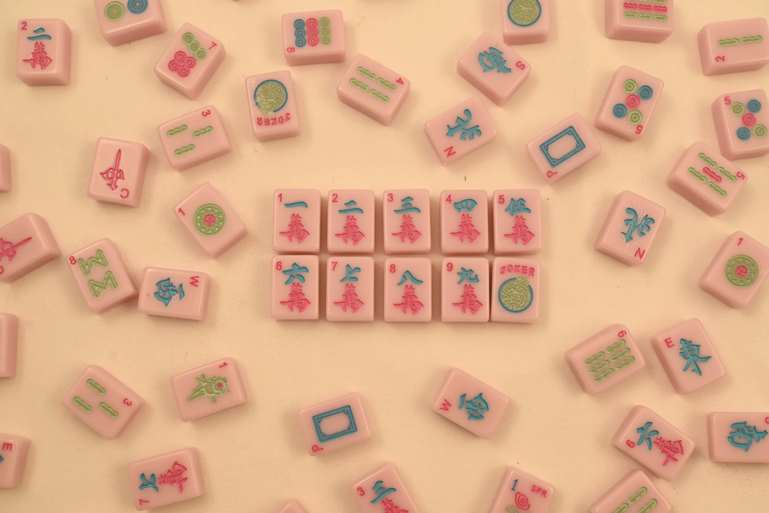 Mahjong tiles with pink backs, arranged on a beige surface, featuring Chinese characters and symbols in red, blue, and green. Some tiles are scattered around, some face up showing their designs.