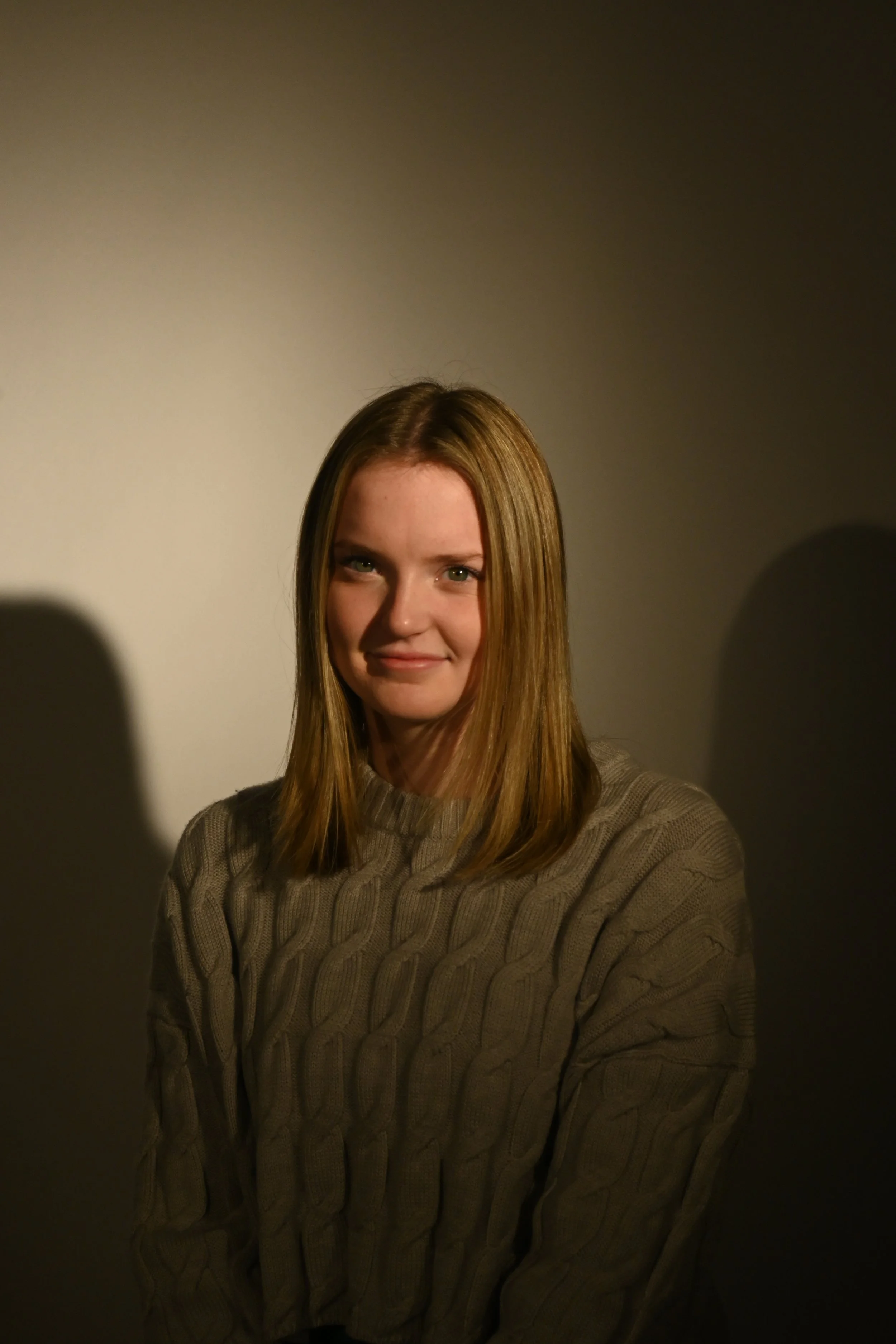 A young woman with shoulder-length straight hair wearing a beige cable-knit sweater, smiling slightly, illuminated by warm lighting against a neutral background.