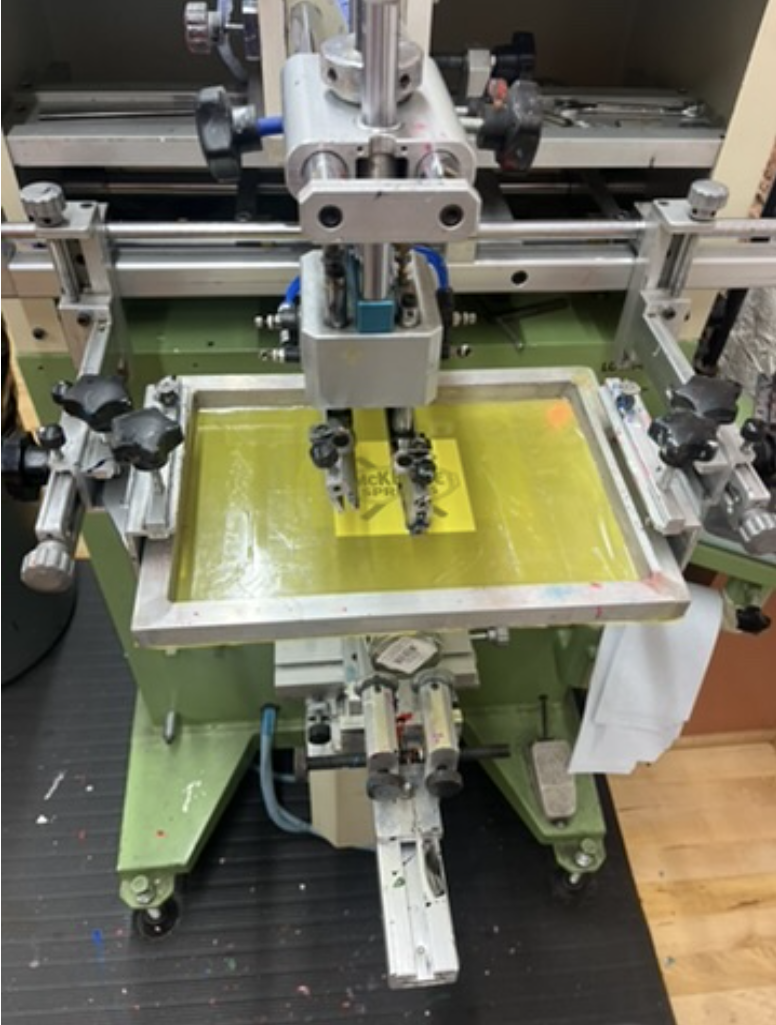 A screen printing machine with a yellow stencil on a green pad, surrounded by multiple adjustable clamps and metal rods.