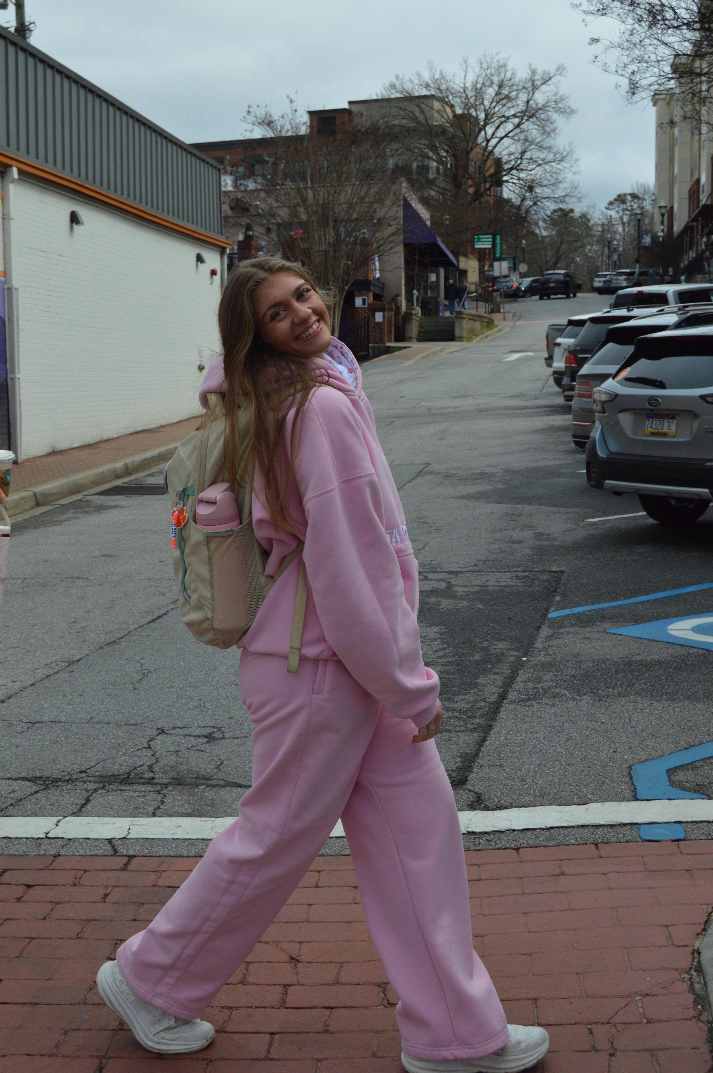 A young woman in a pink hoodie and matching sweatpants, smiling and standing on a crosswalk in a city parking lot, carrying a beige backpack.