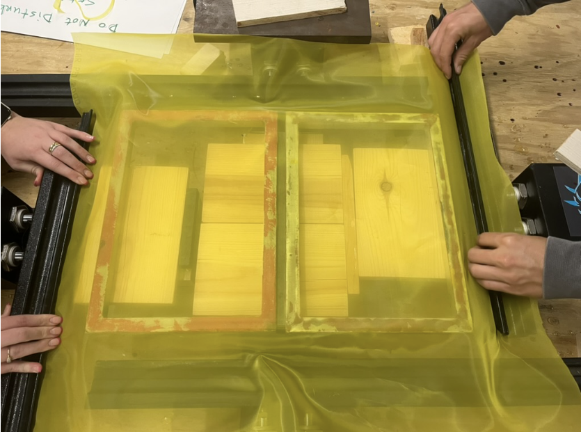 Two people handle a vacuum chamber covering a rectangular glass mold with yellow wooden blocks, likely for a scientific or manufacturing process.