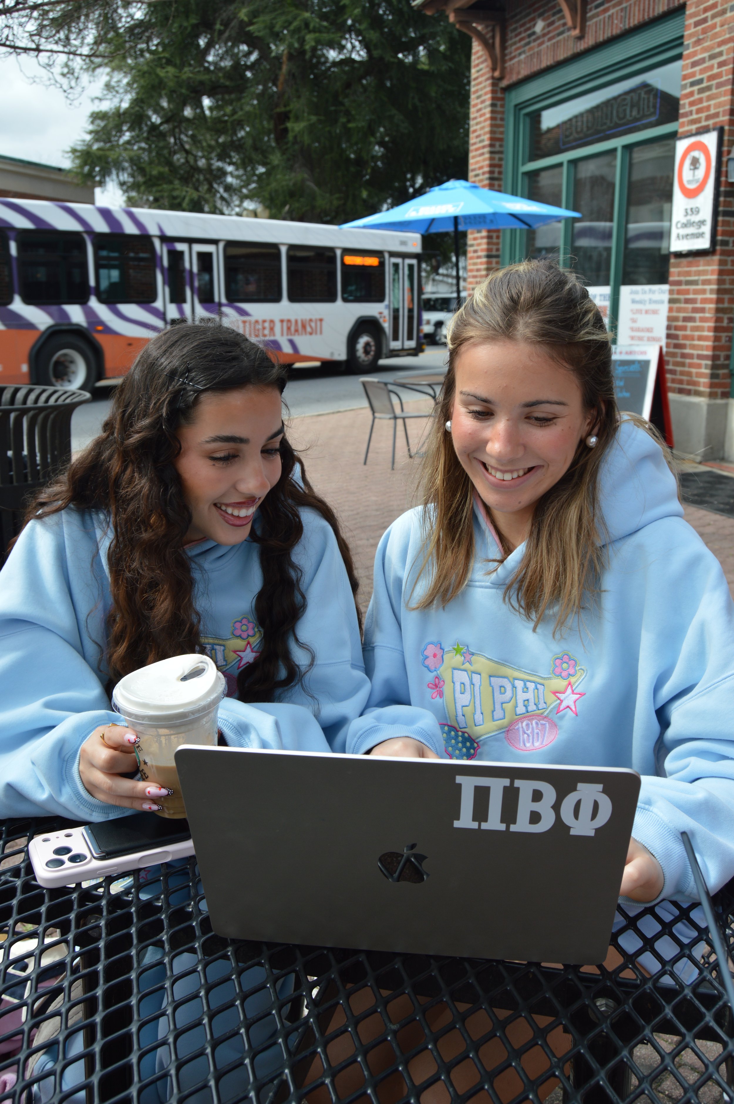Two young women sitting at an outdoor table, smiling and looking at a laptop. One is holding a drink, and both are wearing light blue hoodies with Greek letters and decorative patches. A bus and a building with a sign are visible in the background.