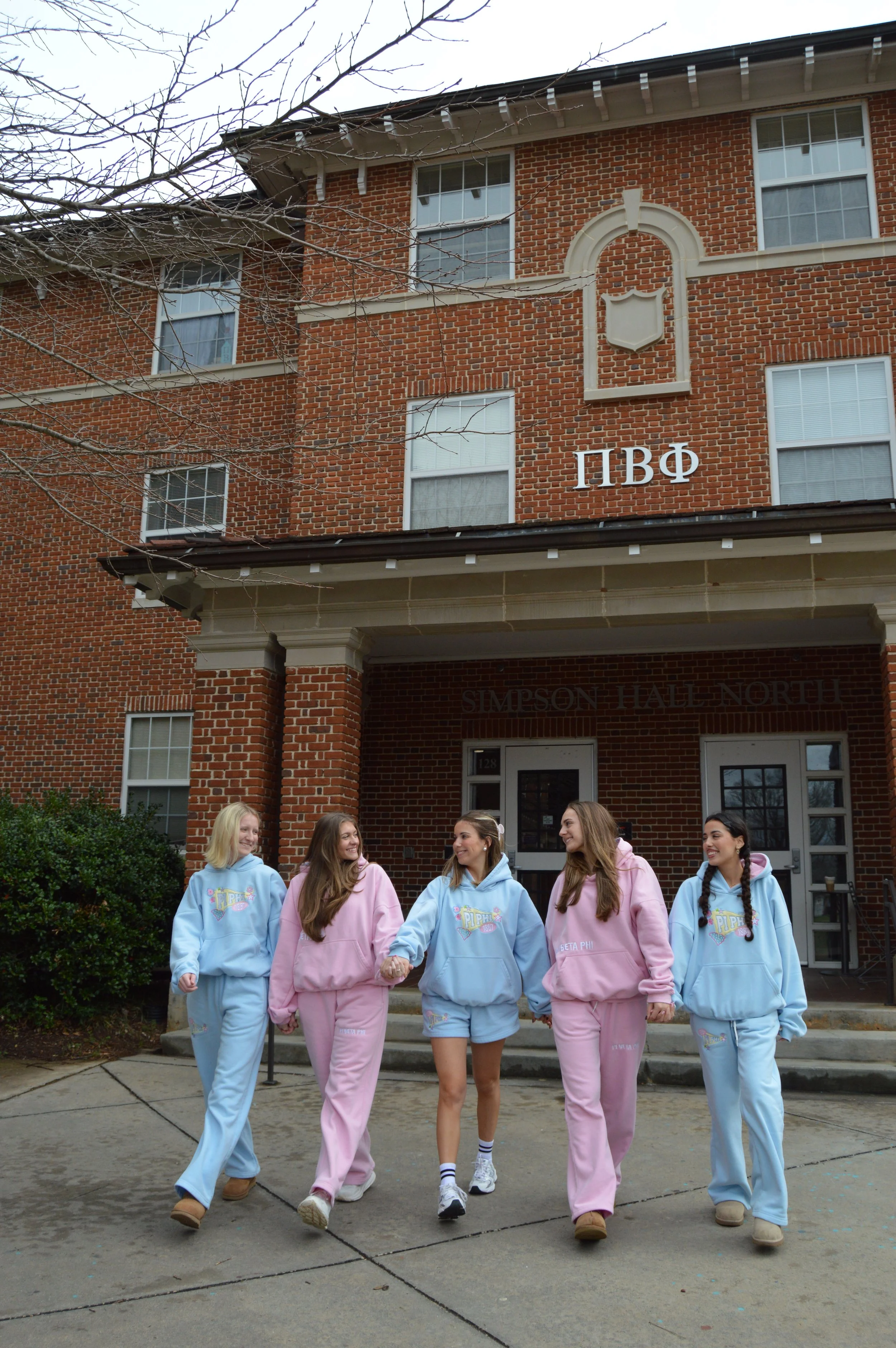 Group of five young women in pastel-colored matching sweatshirts and pants walking hand-in-hand in front of a brick dormitory building on a college campus, with leafless trees and the sign 'SIMPSON HALL NORTH' visible.