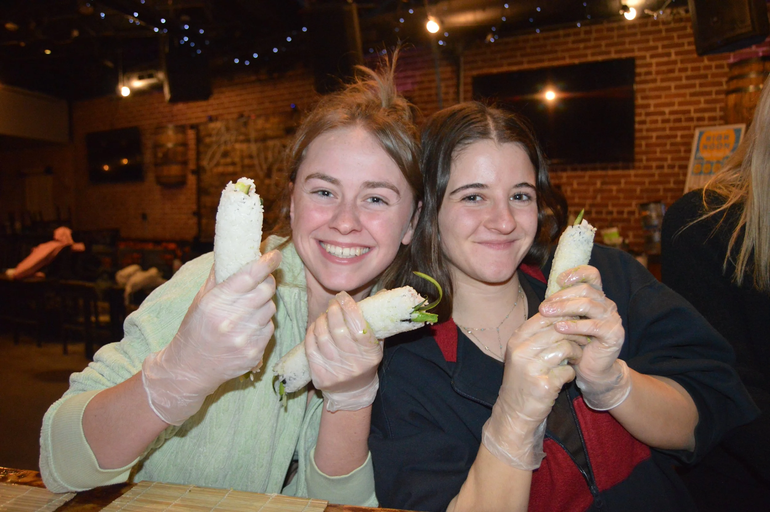 Two women smiling and holding wrapped sandwiches inside a restaurant with brick walls and television screens.