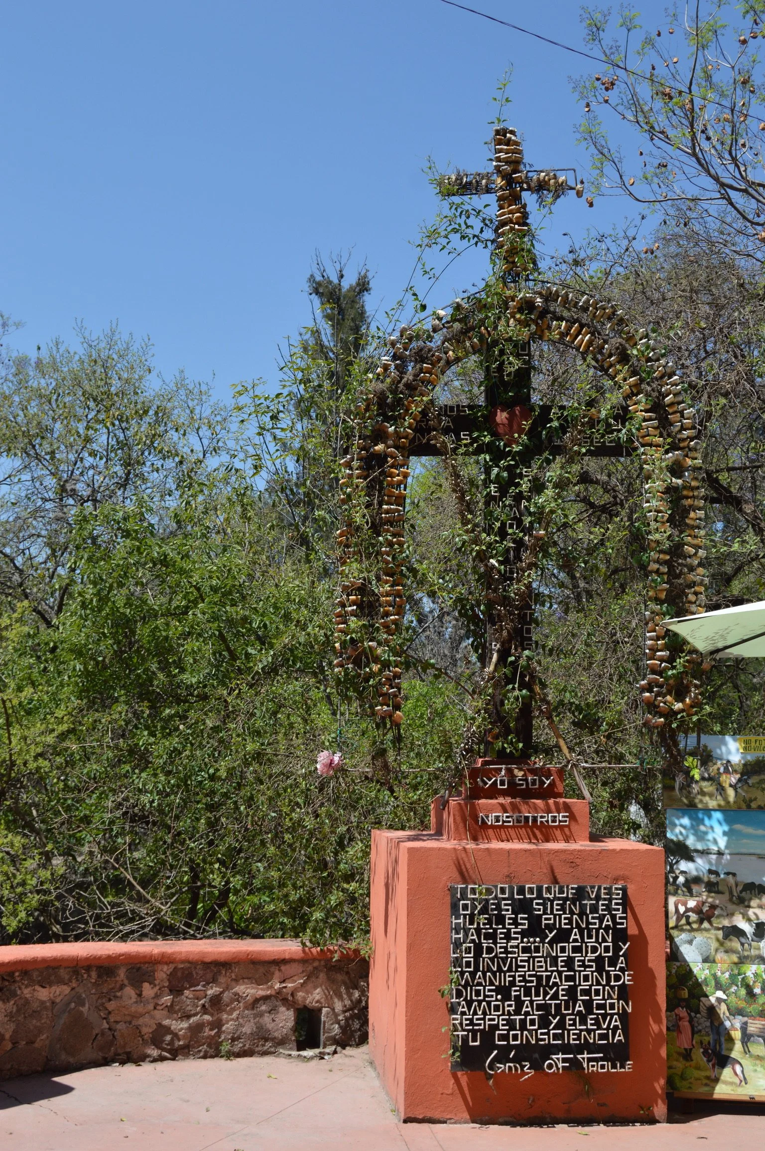 A large cross made of wood and decorated with numerous small objects, situated outdoors against a background of trees and a clear blue sky. There is a pink flower attached to the cross, and a plaque with text in Spanish in front of the display.