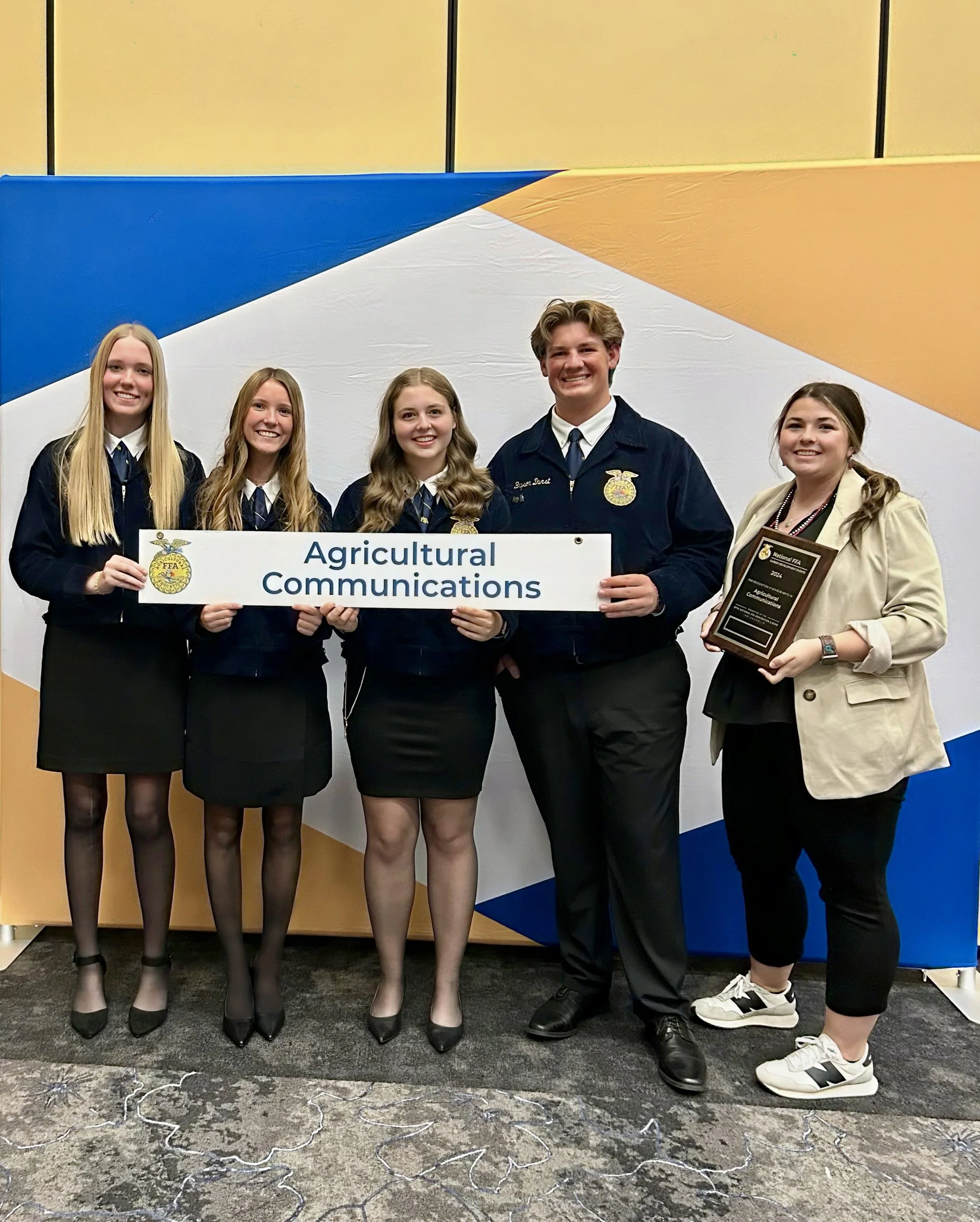Group of five teenagers dressed in FFA jackets holding a sign that reads 'Agricultural Communications', with one person holding an award plaque, standing in front of a colorful geometric backdrop.