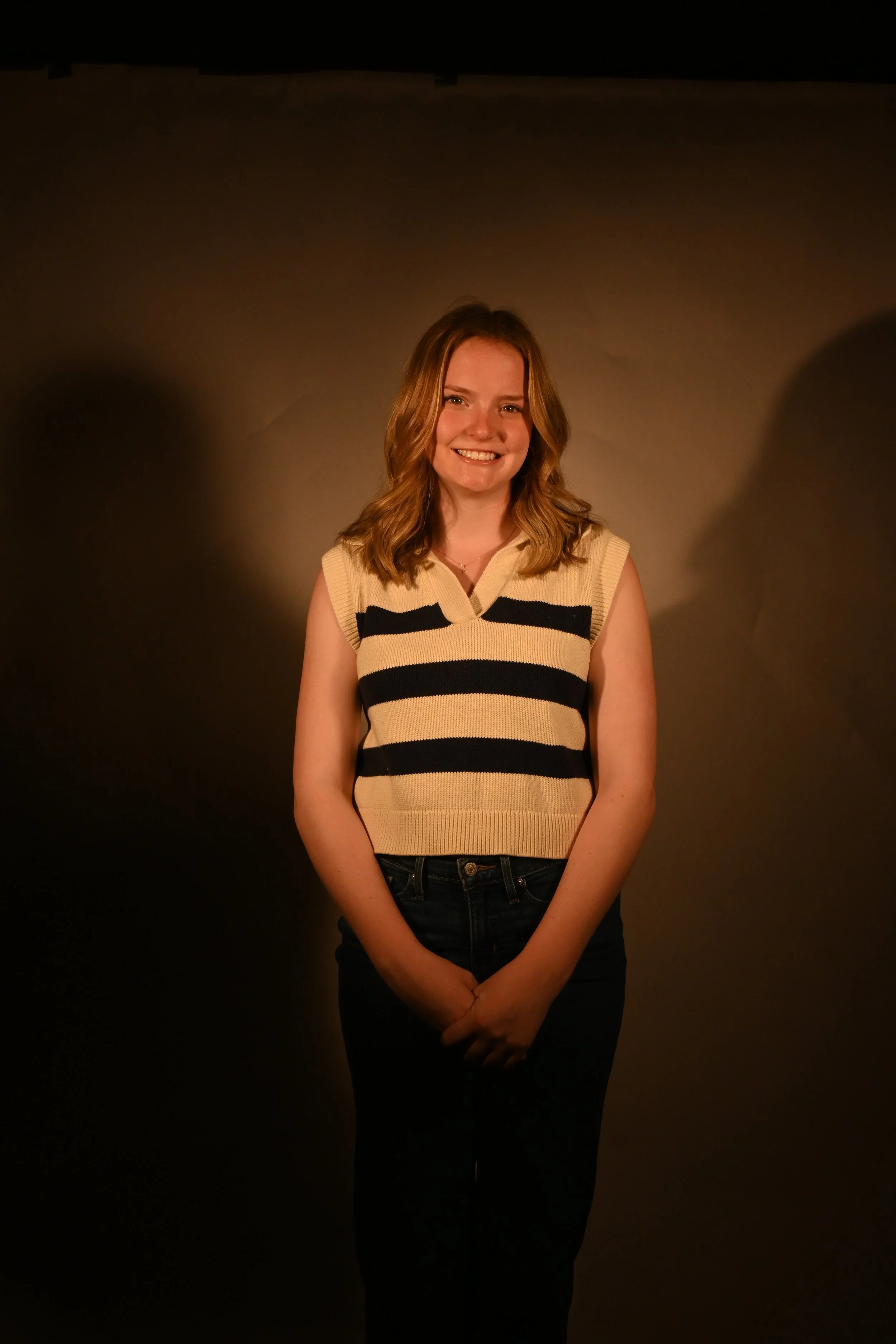 Young woman with shoulder-length wavy hair, smiling, wearing a sleeveless beige and black striped sweater, standing against a dark background with subtle lighting.