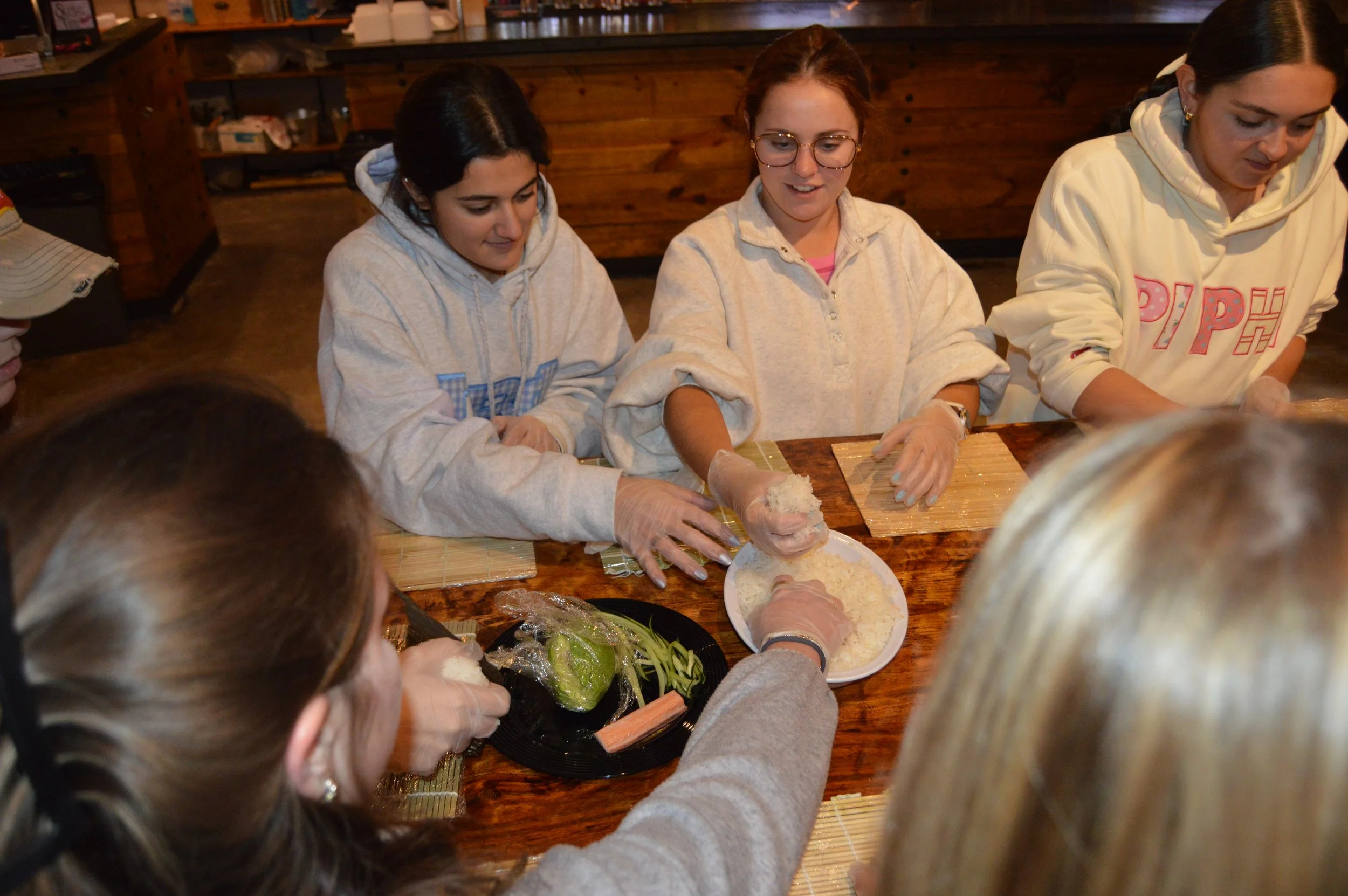 Group of women participating in a sushi-making class, assembling sushi rolls with rice, vegetables, and seafood on a wooden table.