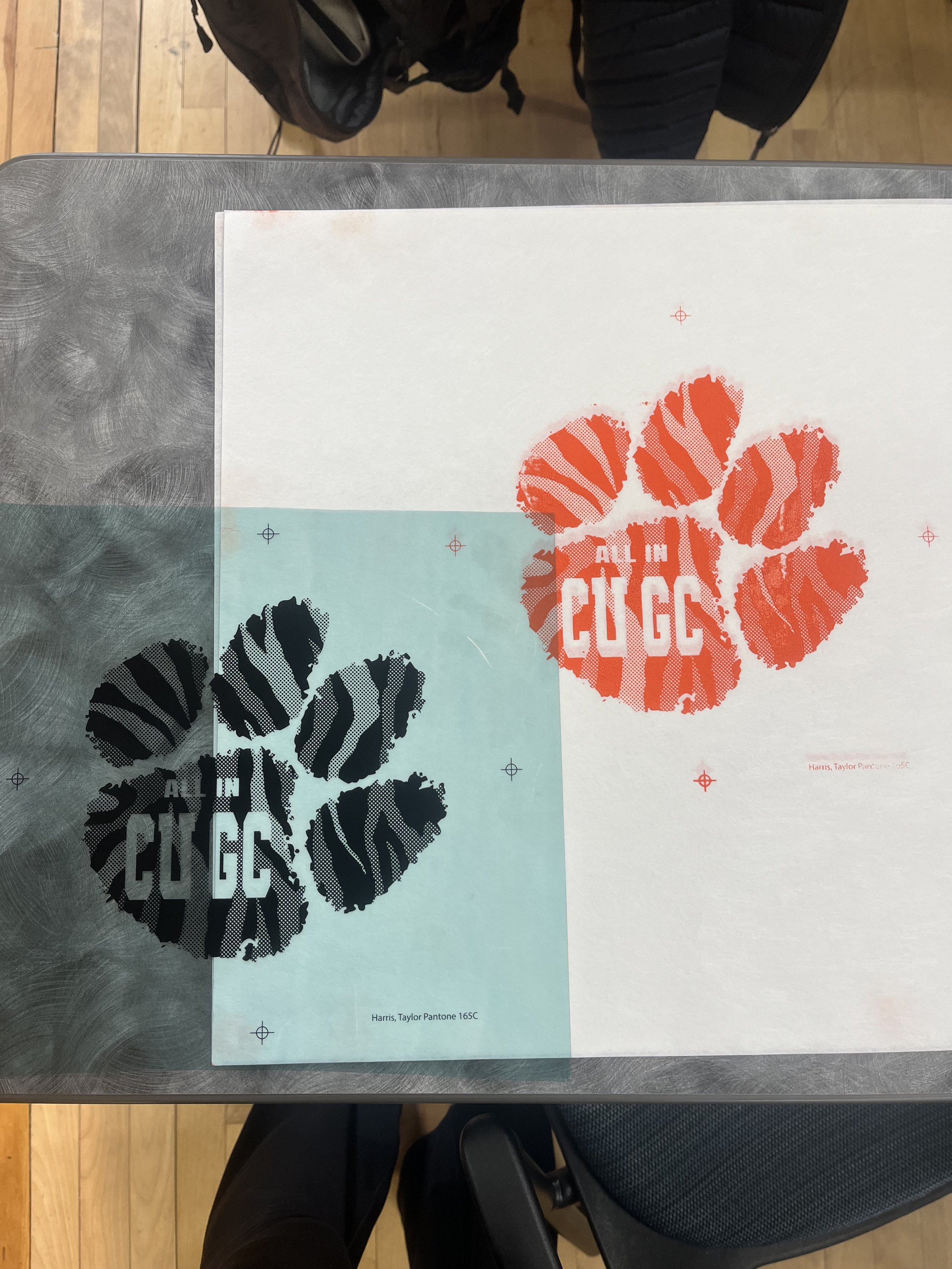 Two prints of a paw print with the phrase 'ALL IN CUGG' in the center, one in black and one in red, placed on a table with a gray surface. The prints appear to be part of an art or design project.