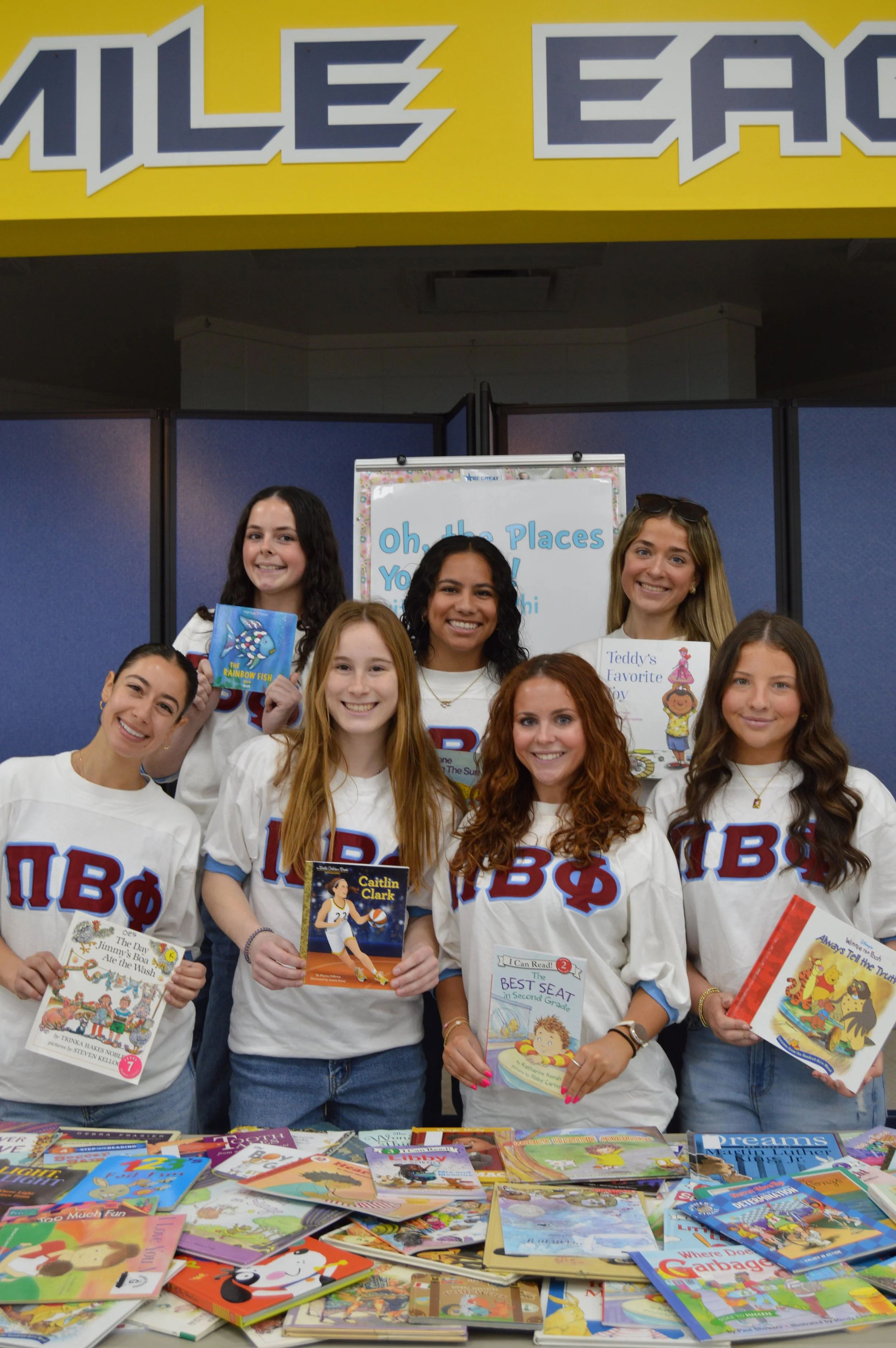 Group of women wearing white T-shirts with Greek letters, standing behind a table full of children's books, at a book fair or signing event.