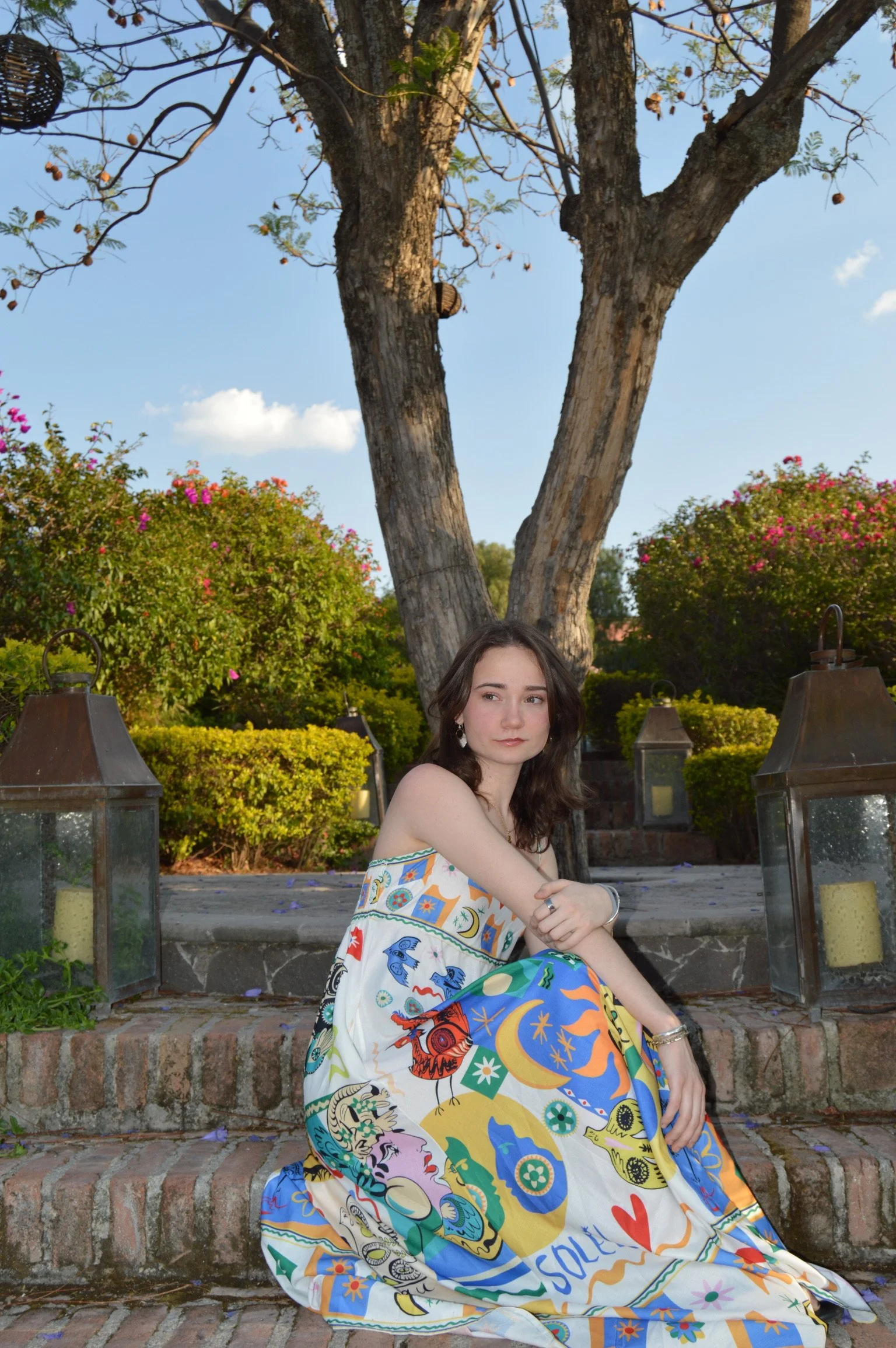 A young woman with dark brown hair sitting on brick steps outdoors. She is wearing a colorful, patterned dress and looking slightly to her left. Behind her, there is a large tree with a few leaves on the branches, and bright bushes with pink and purple flowers. On each side of her, there are vintage-style lanterns with candles inside.