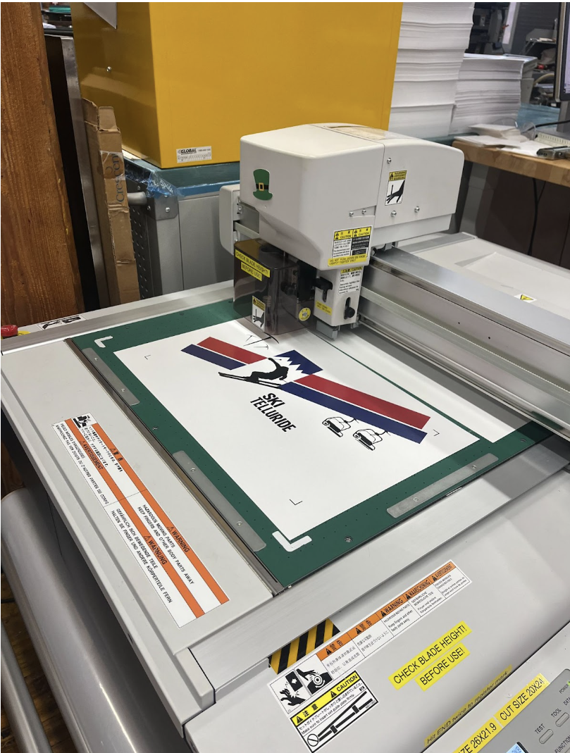 A flatbed UV printer printing a graphic design on a white surface, with various warning labels and a green hat sticker on the printer, in an industrial workspace.