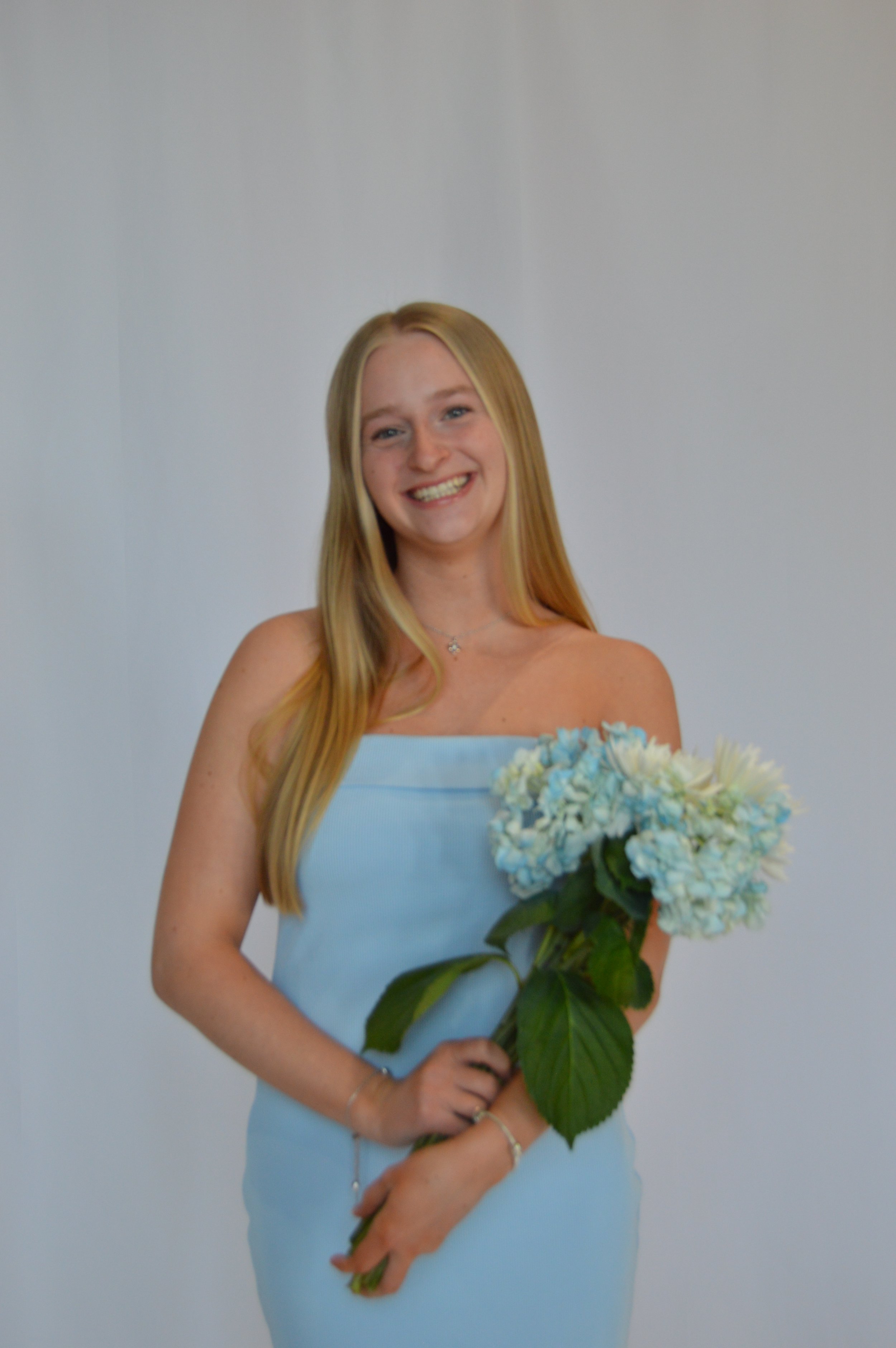 A young woman with long blonde hair smiling, wearing a light blue strapless dress, holding a bouquet of white and blue flowers, standing against a plain white wall.