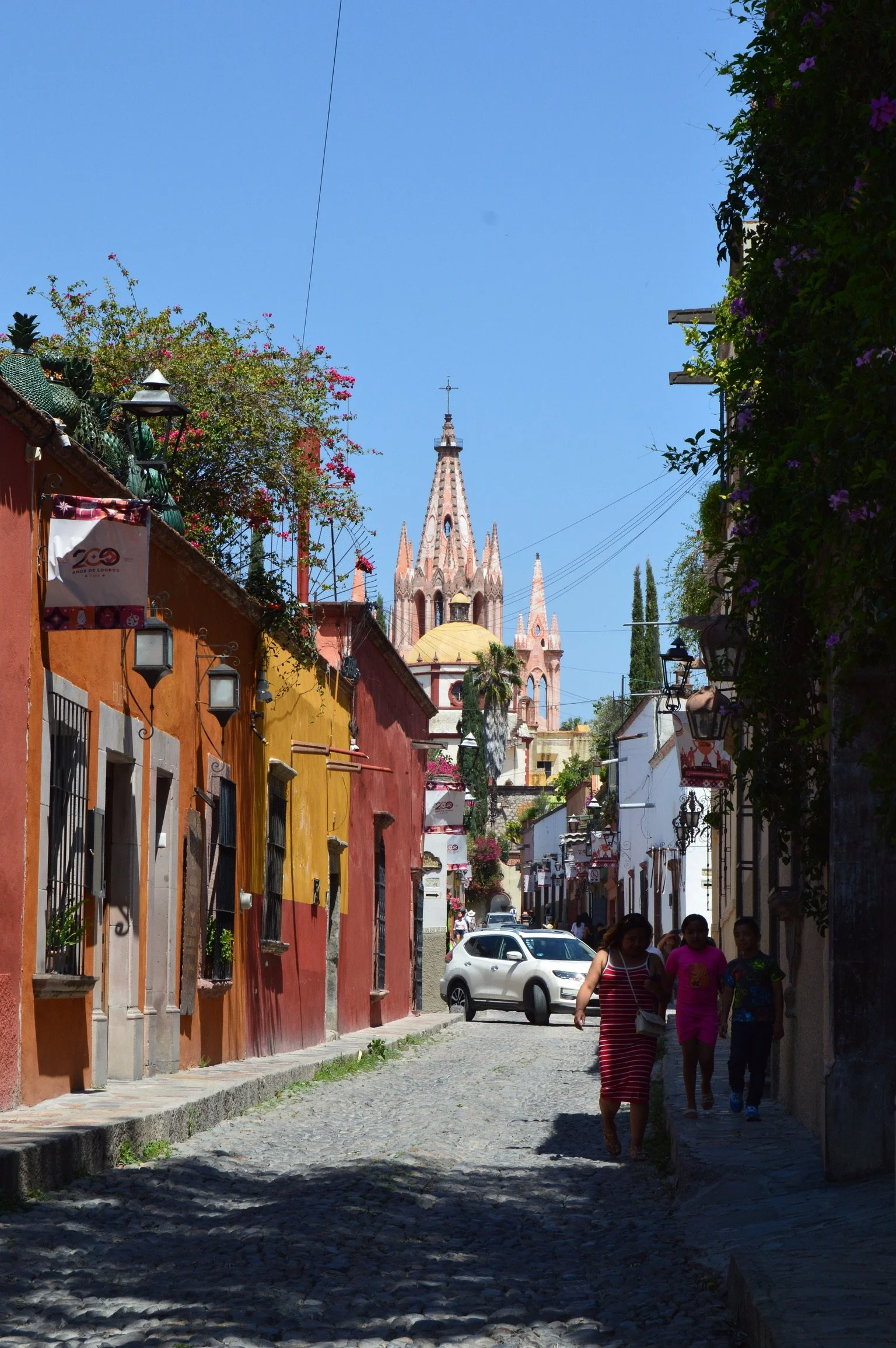 Colorful street with cobblestone path, vibrant buildings, flowers, and a church with a tall spire in the background.