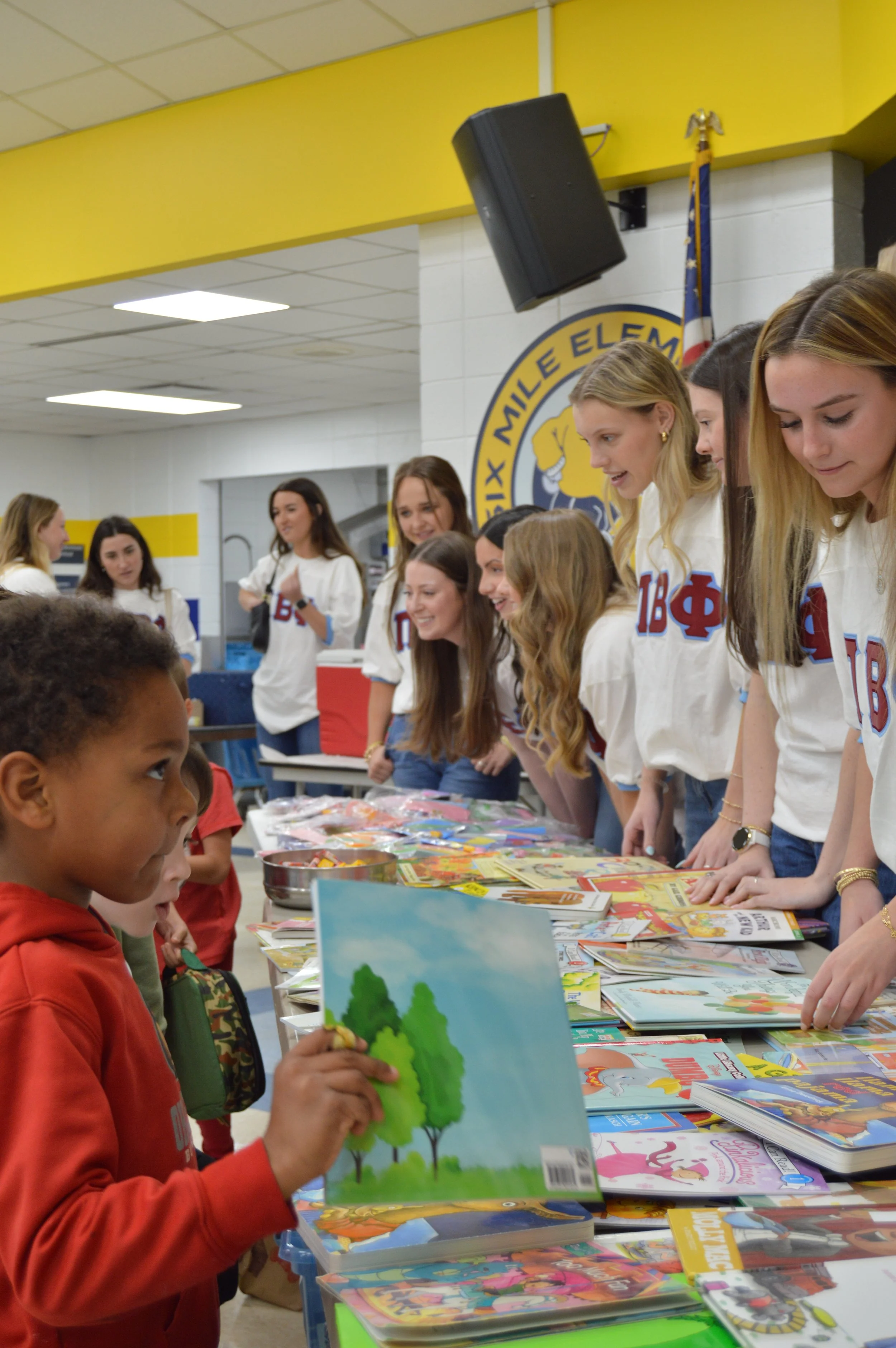 A group of young women wearing white shirts with Greek letters are standing behind tables filled with books and magazines, talking to children in a school cafeteria.