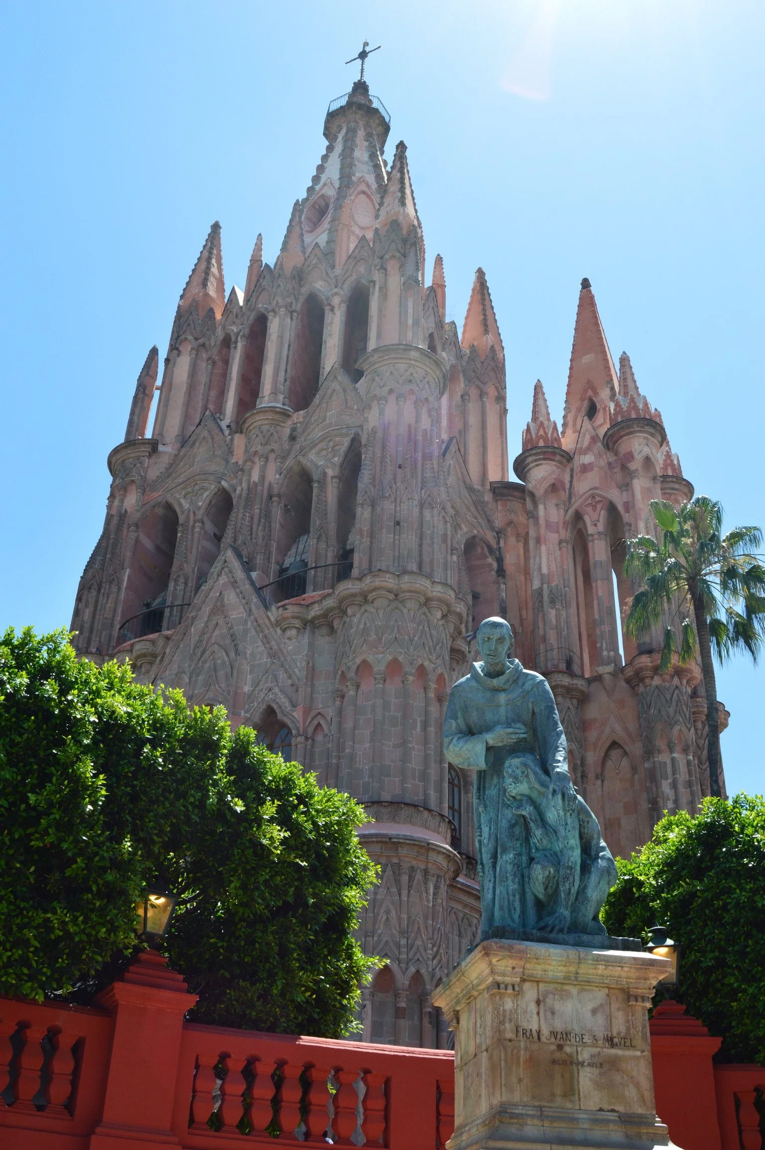 Photo of a pink Gothic-style cathedral with a statue of Fray Juan de S. McKel in the foreground, surrounded by green trees and a bright blue sky.