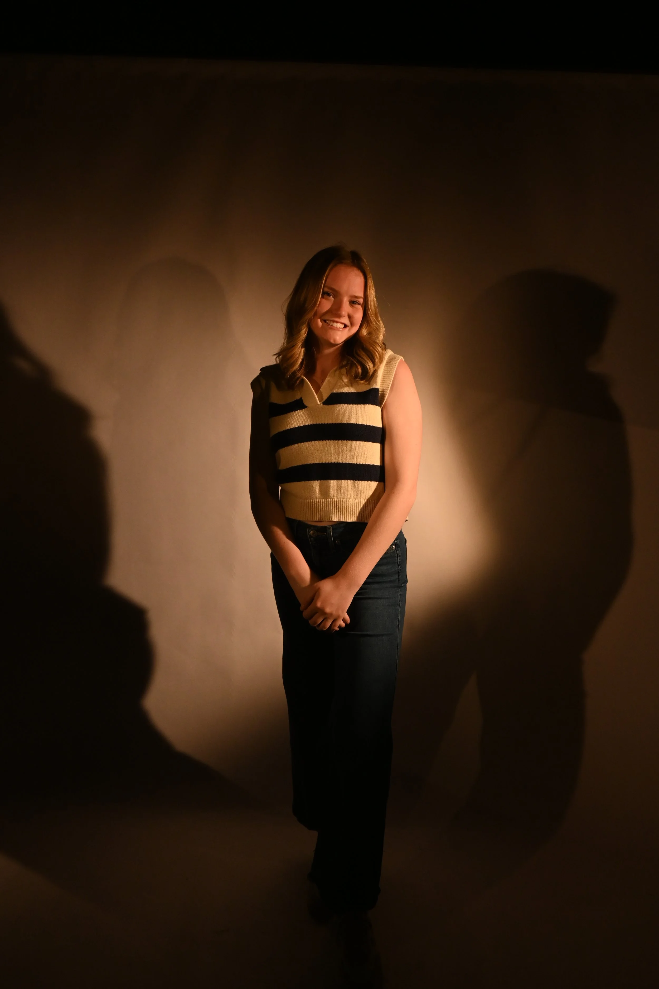 A young woman smiling and standing against a neutral background with shadows cast on the wall behind her.