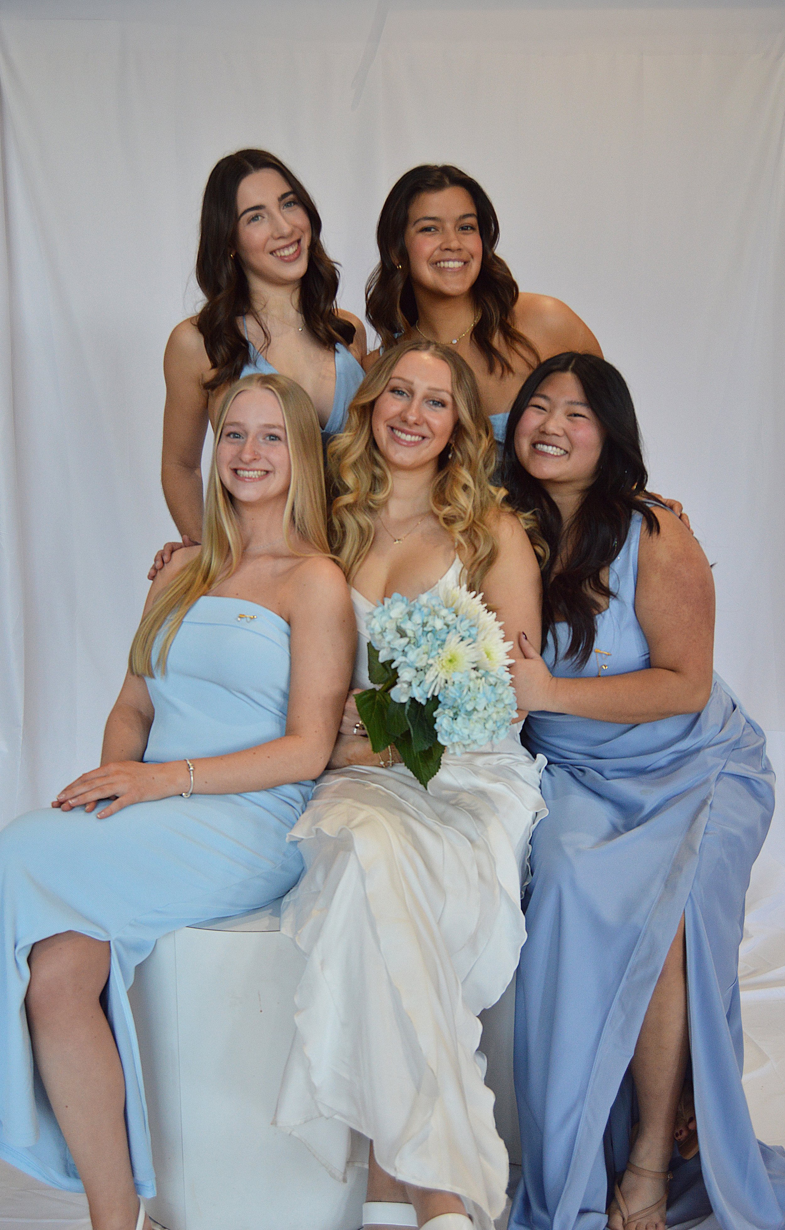 Group of five young women in formal dresses, with the woman in the white dress holding a blue and white bouquet of flowers, seated and smiling in front of a white backdrop.