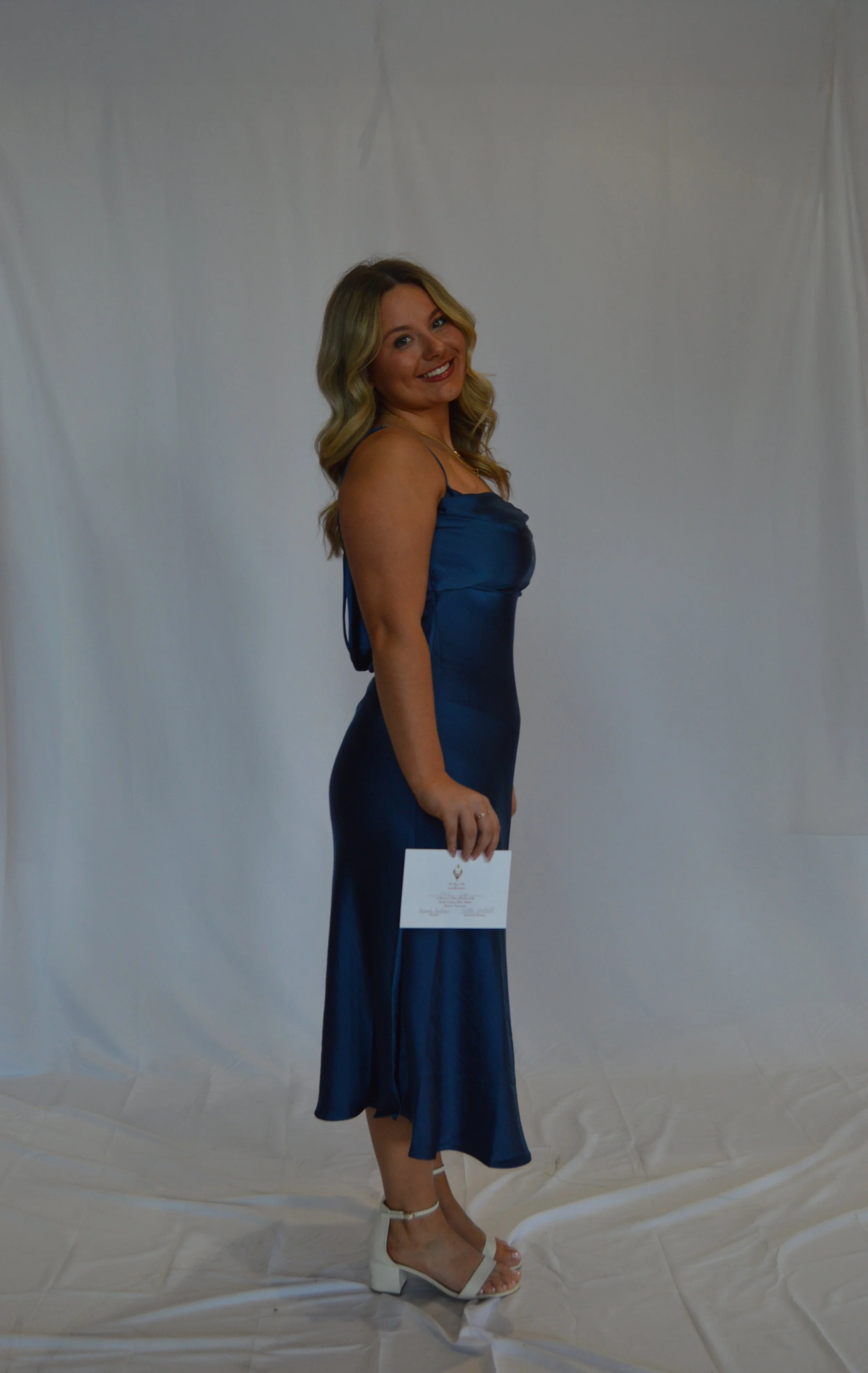 A woman in a blue dress standing against a plain white background, holding a certificate and smiling at the camera.