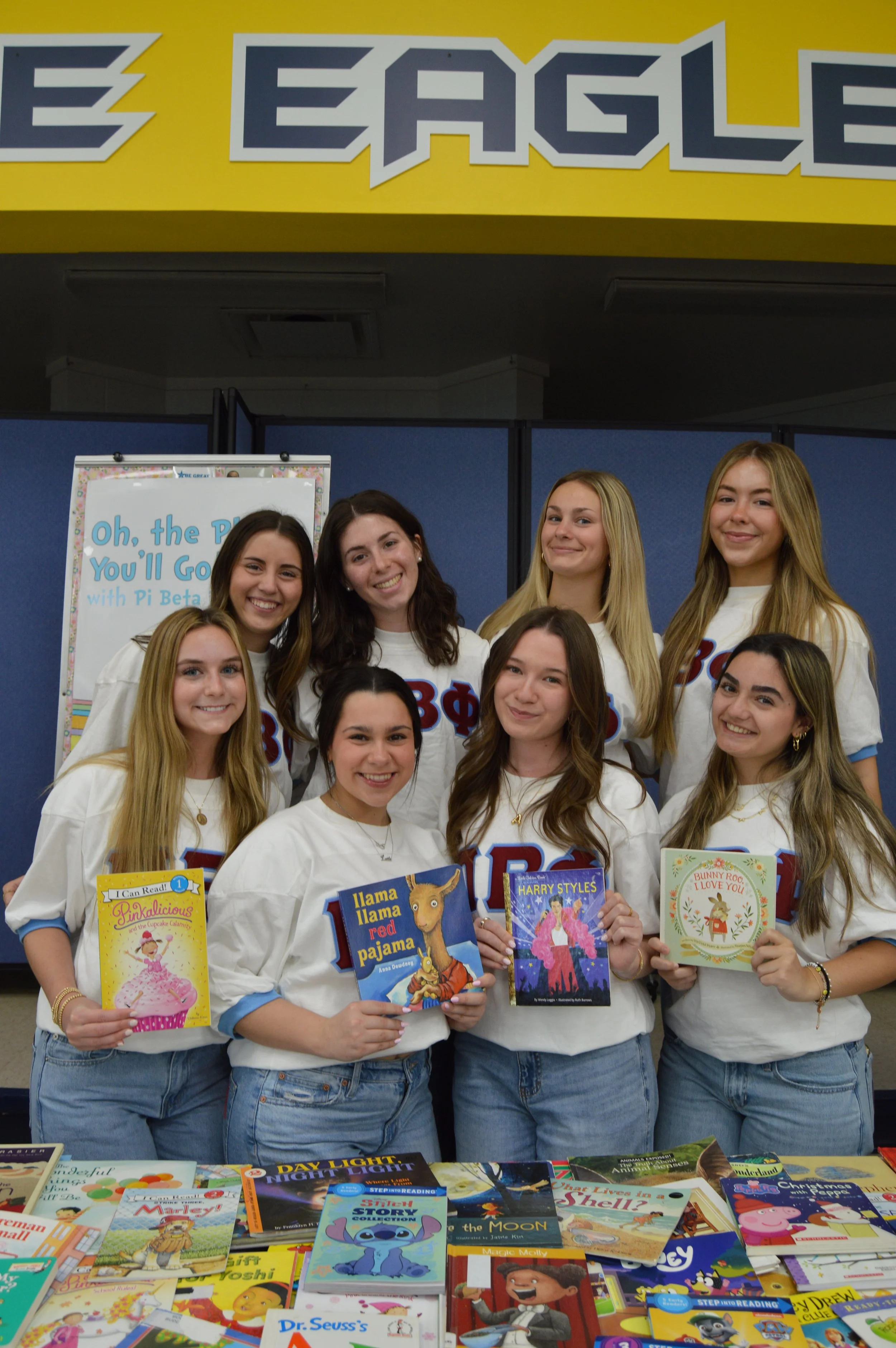 A group of nine young women, all wearing white t-shirts with Greek letters, standing behind a table full of books, smiling for a photo at a bookstore or library event.