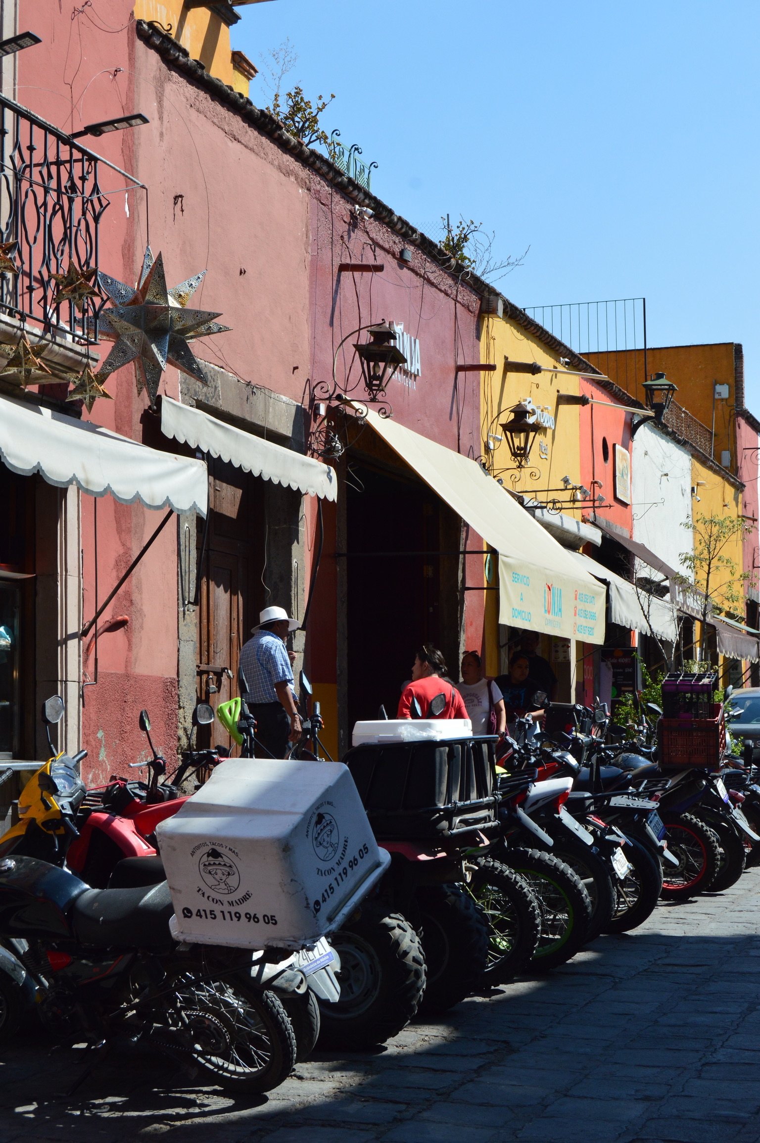 Colorful street scene with motorbikes parked in front of cafes with umbrellas, people engaged in conversation, and vibrant buildings in pink, yellow, orange, and white under a clear blue sky.