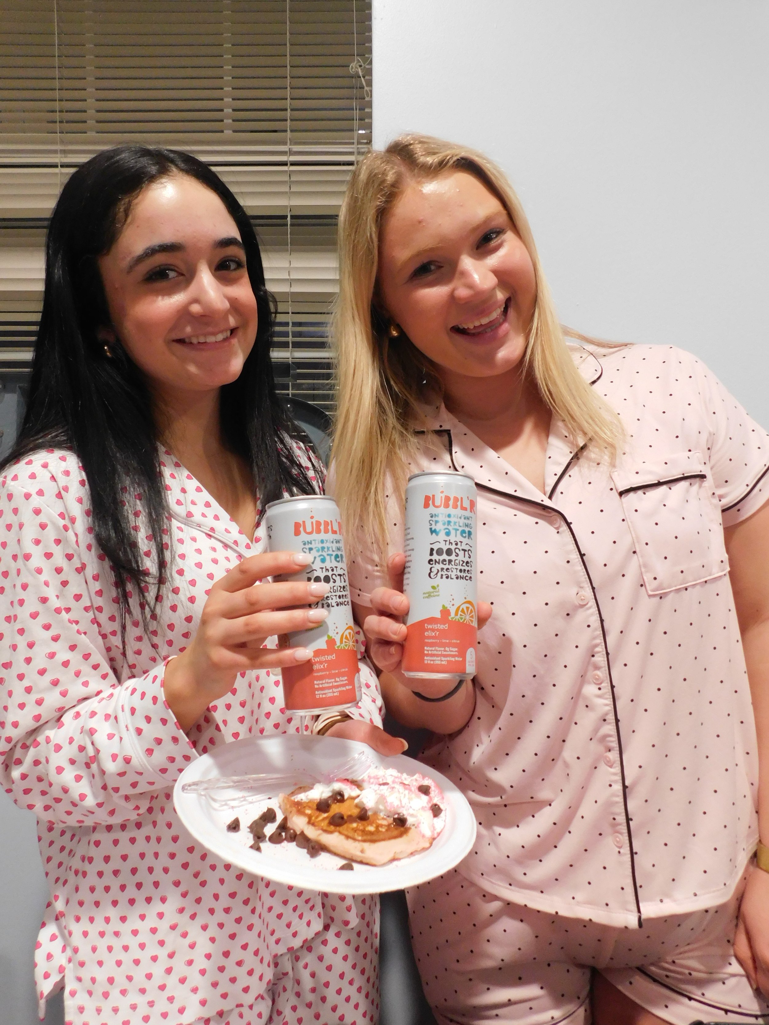 Two women wearing pink and white pajamas with polka dots, holding cans of sparkling water, standing indoors next to a window with blinds, with a plate of decorated cake or pie.